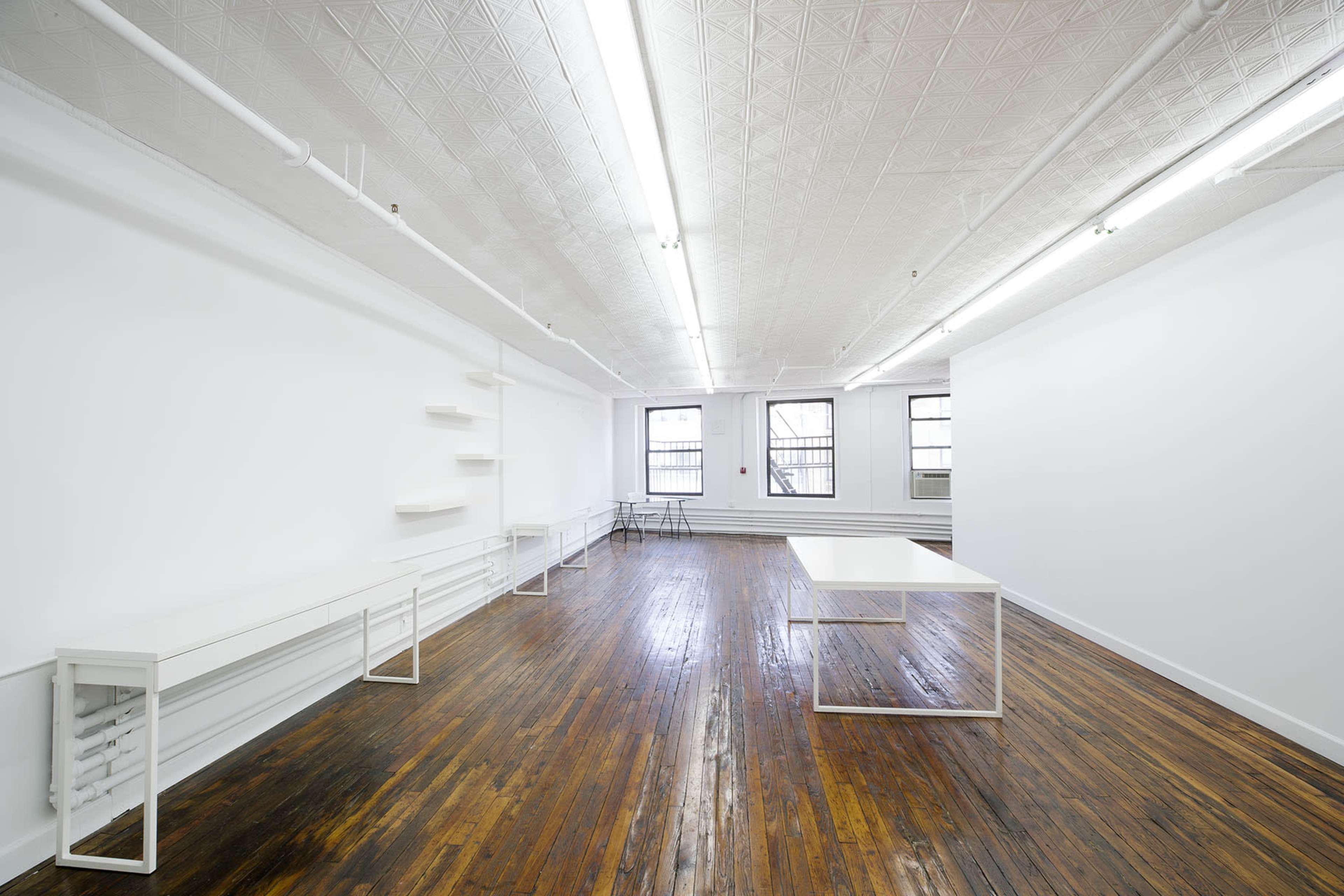 The image shows a spacious, minimalistic room with a hardwood floor, white walls, several empty shelves, and a large white table at the center.