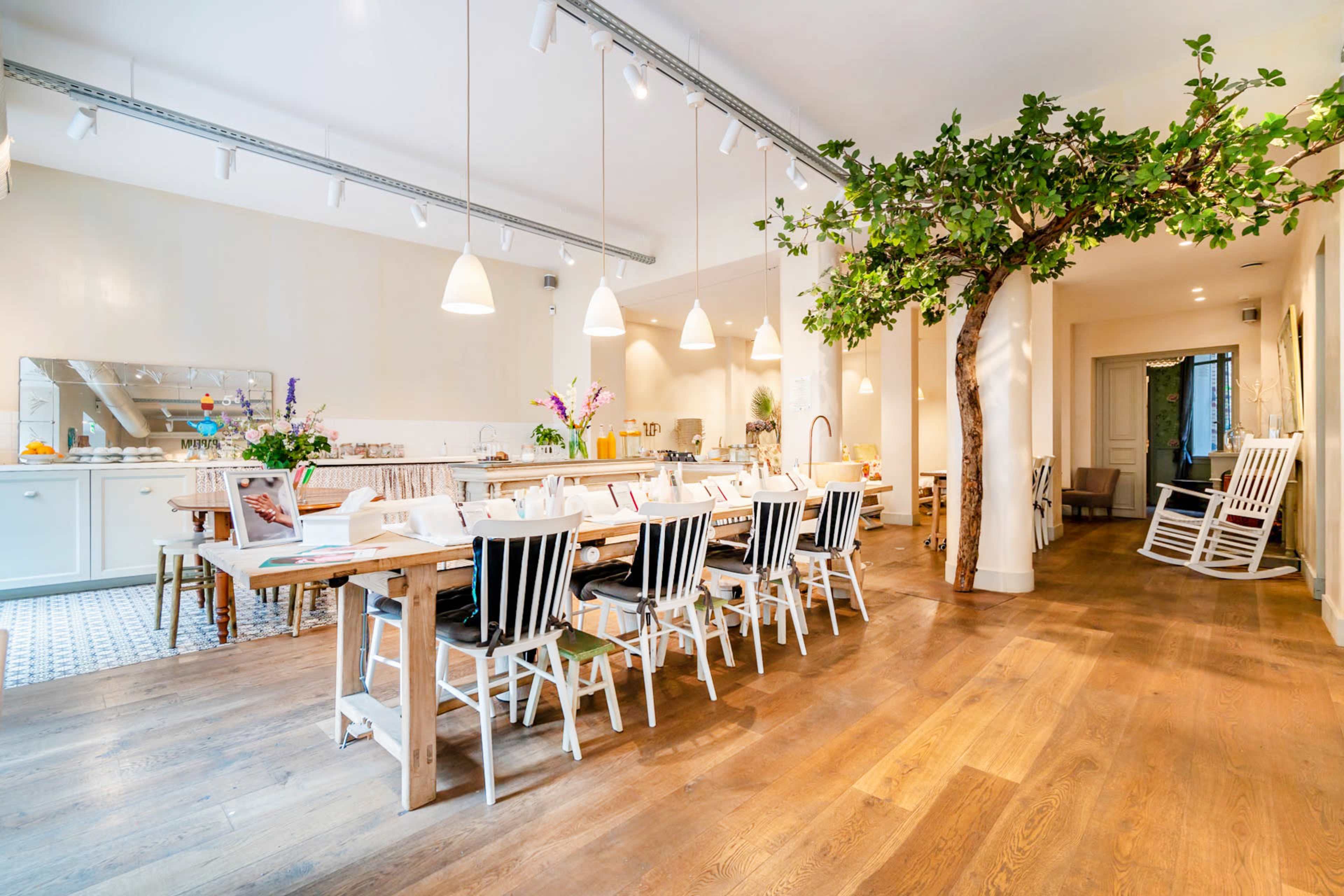 A spacious dining area with a long wooden table surrounded by black and white chairs, featuring a large indoor tree and modern pendant lighting.
