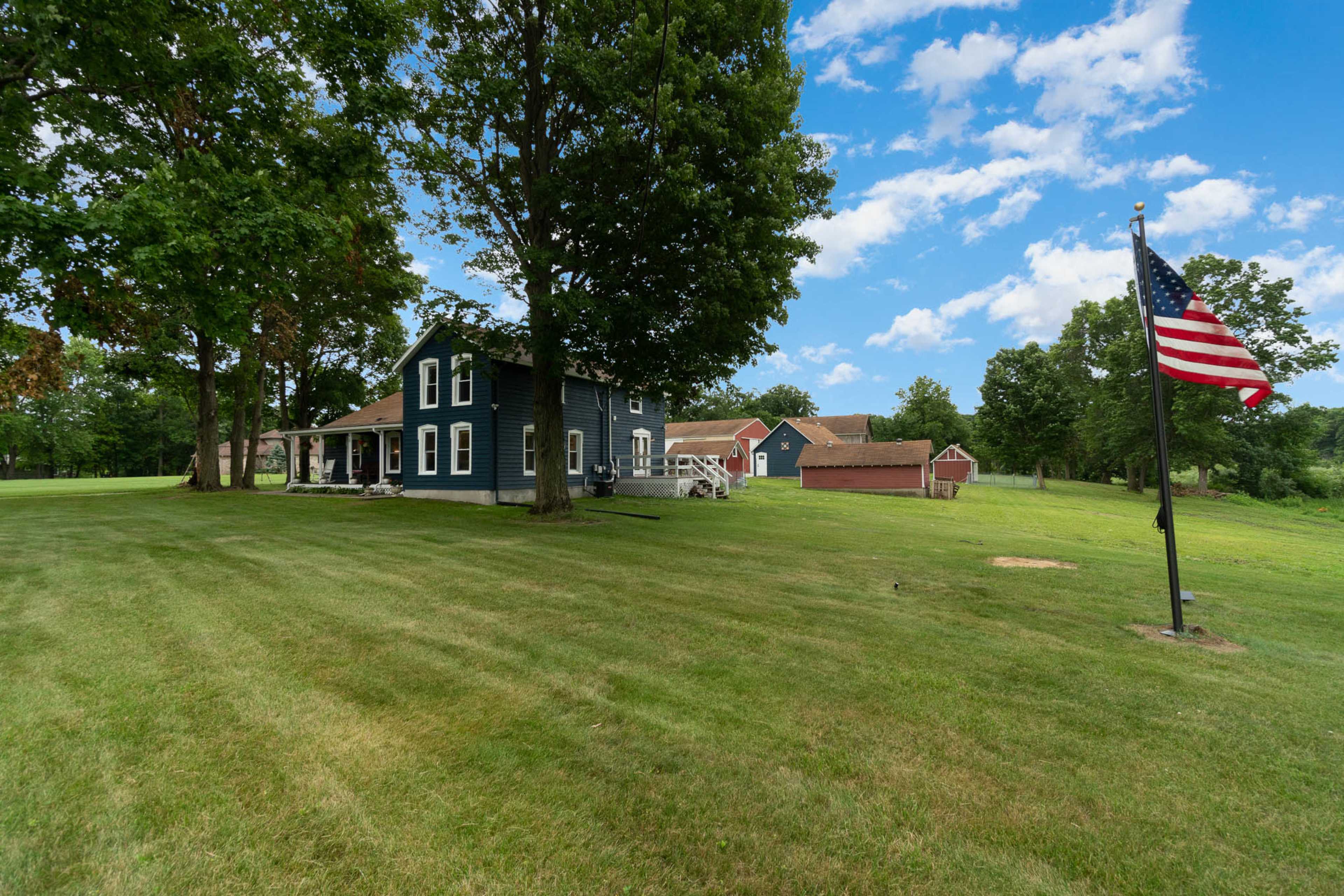 A blue two-story house with a porch, surrounded by green grass and trees, is situated near a red barn and flying an American flag.