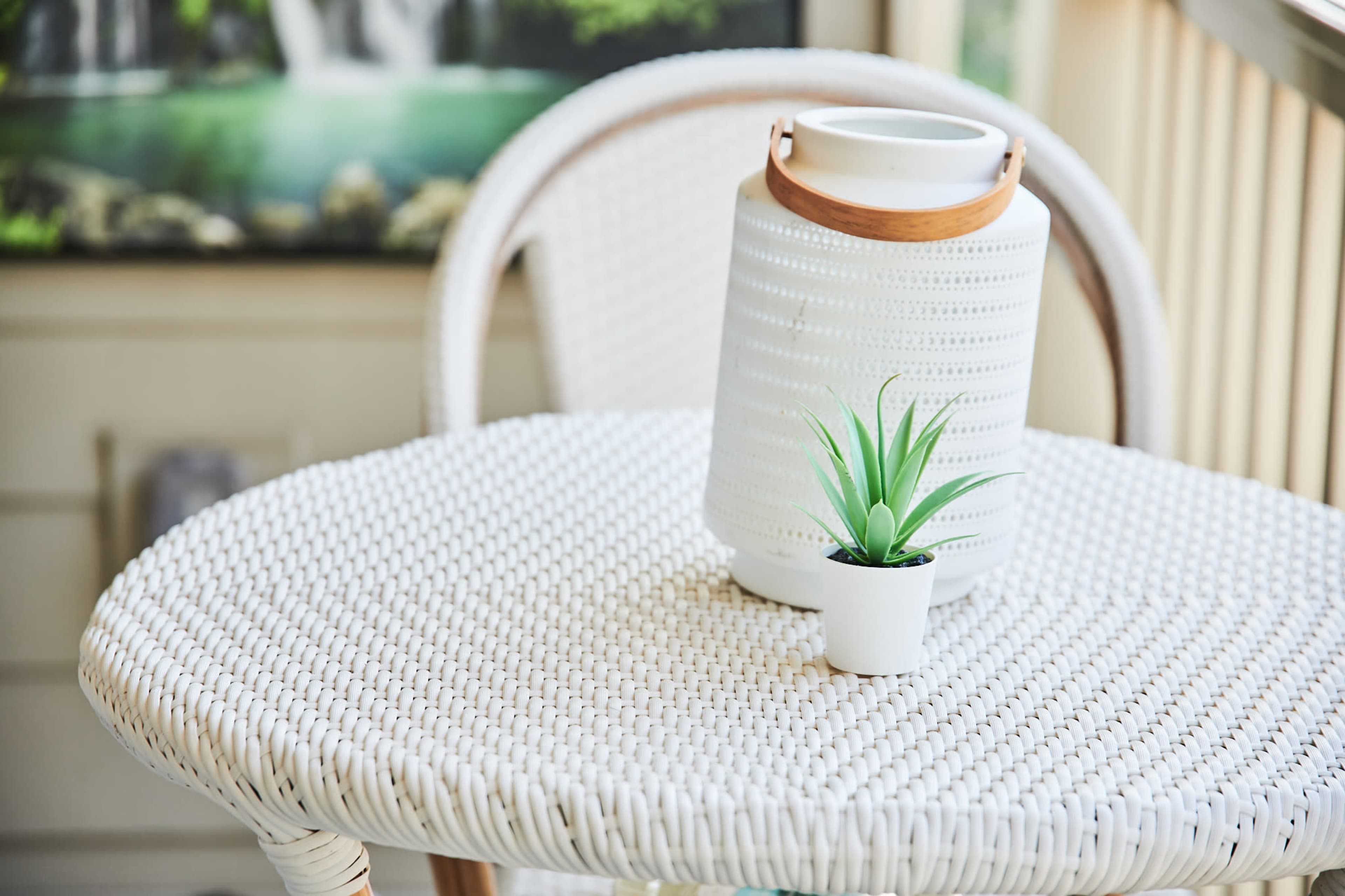 A white woven table holds a small potted plant and a lantern on a porch with a landscape painting in the background.