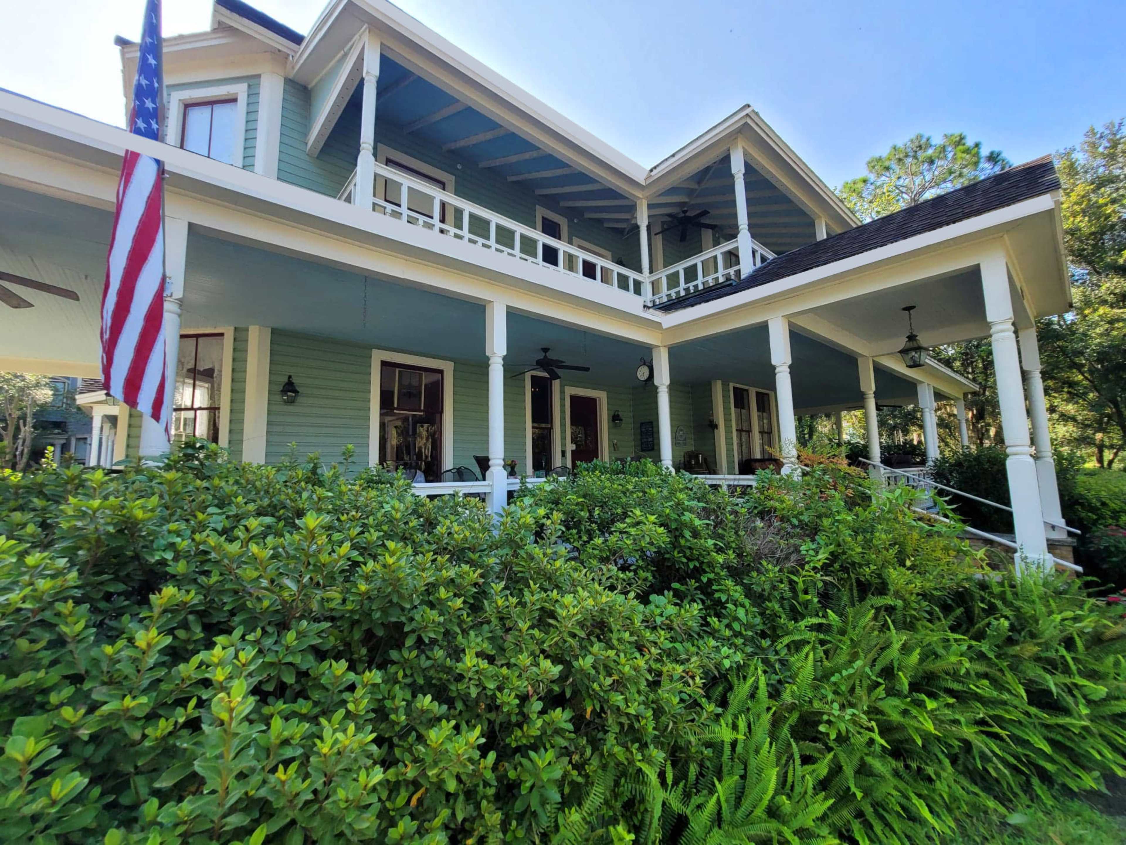 The image shows a two-story house with a wraparound porch, surrounded by greenery and featuring an American flag on the left side.