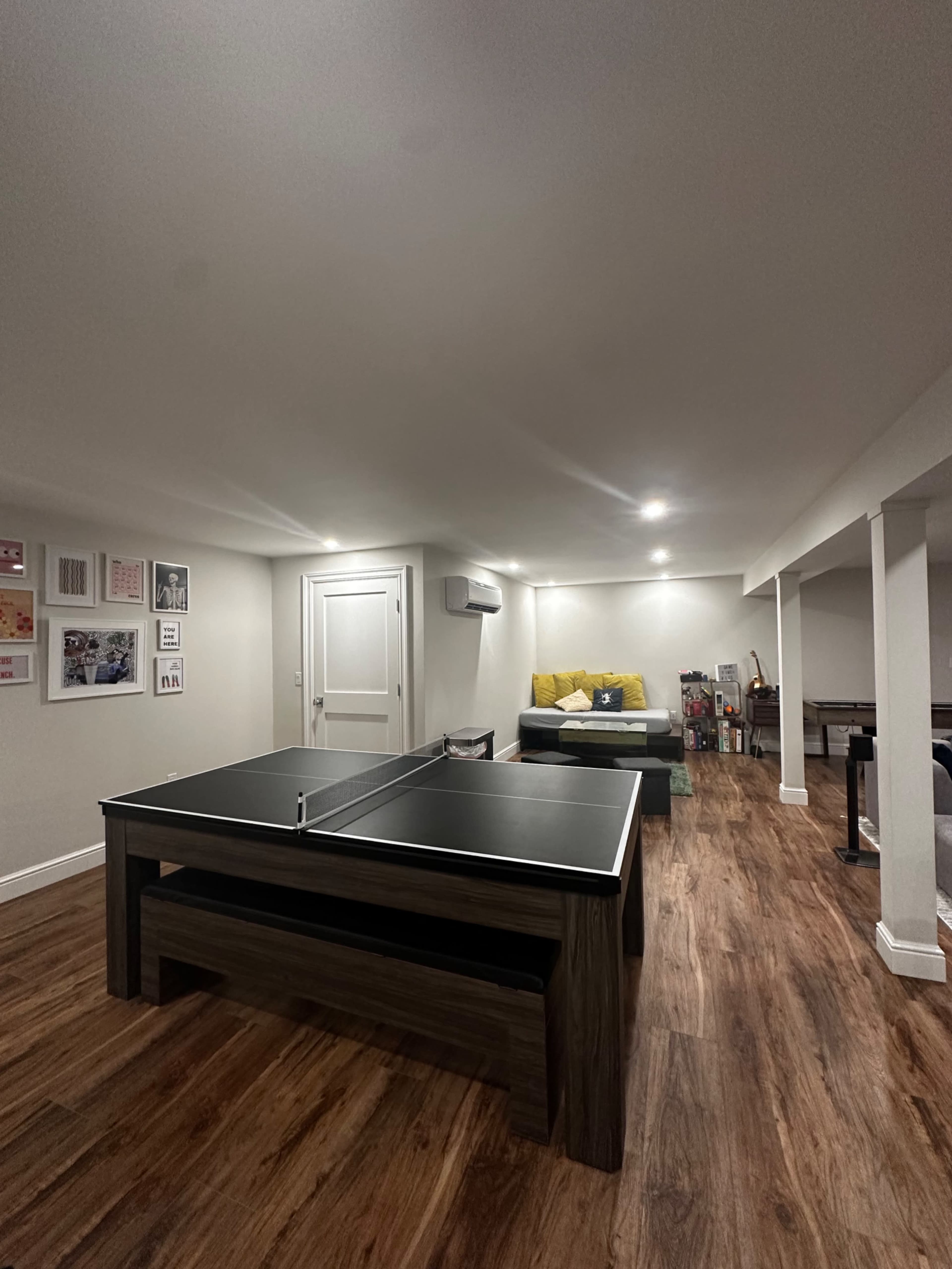 A basement recreation room with a ping pong table in the center and a couch in the background.