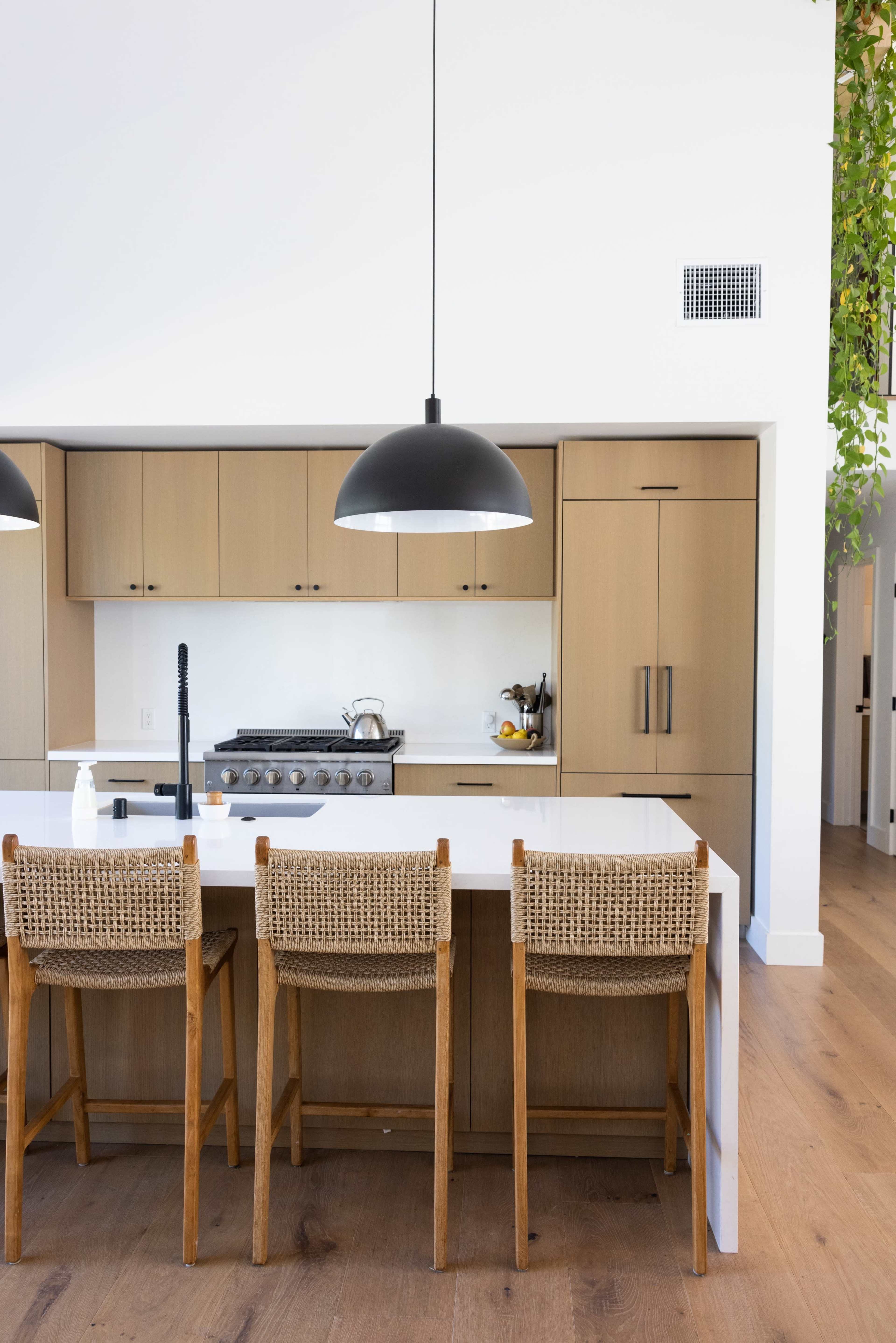 A modern kitchen features a large island with three wooden bar stools, a black pendant light overhead, and beige cabinetry.