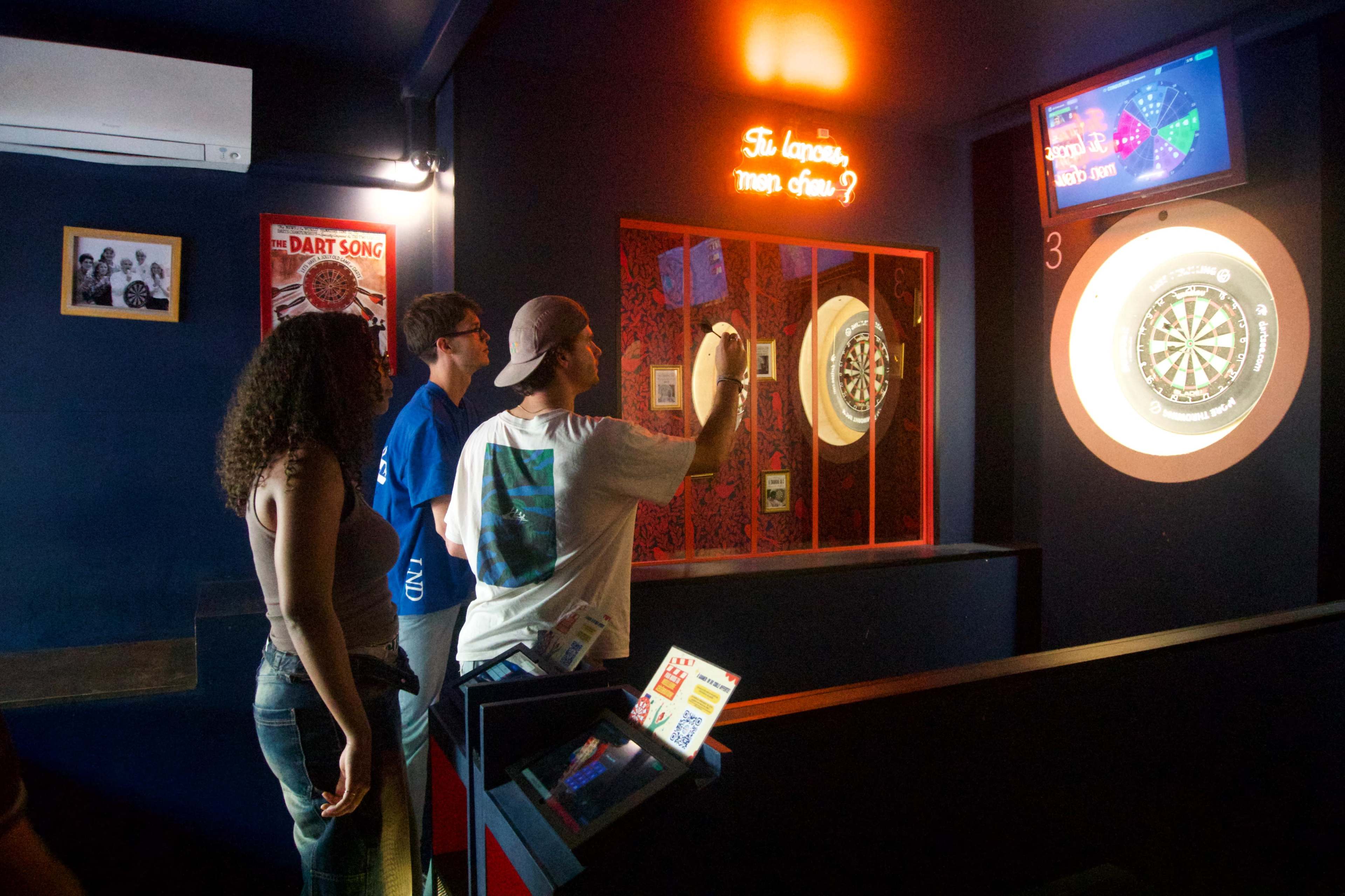Three people are playing darts in a dimly lit room featuring dart boards and a digital scoreboard.