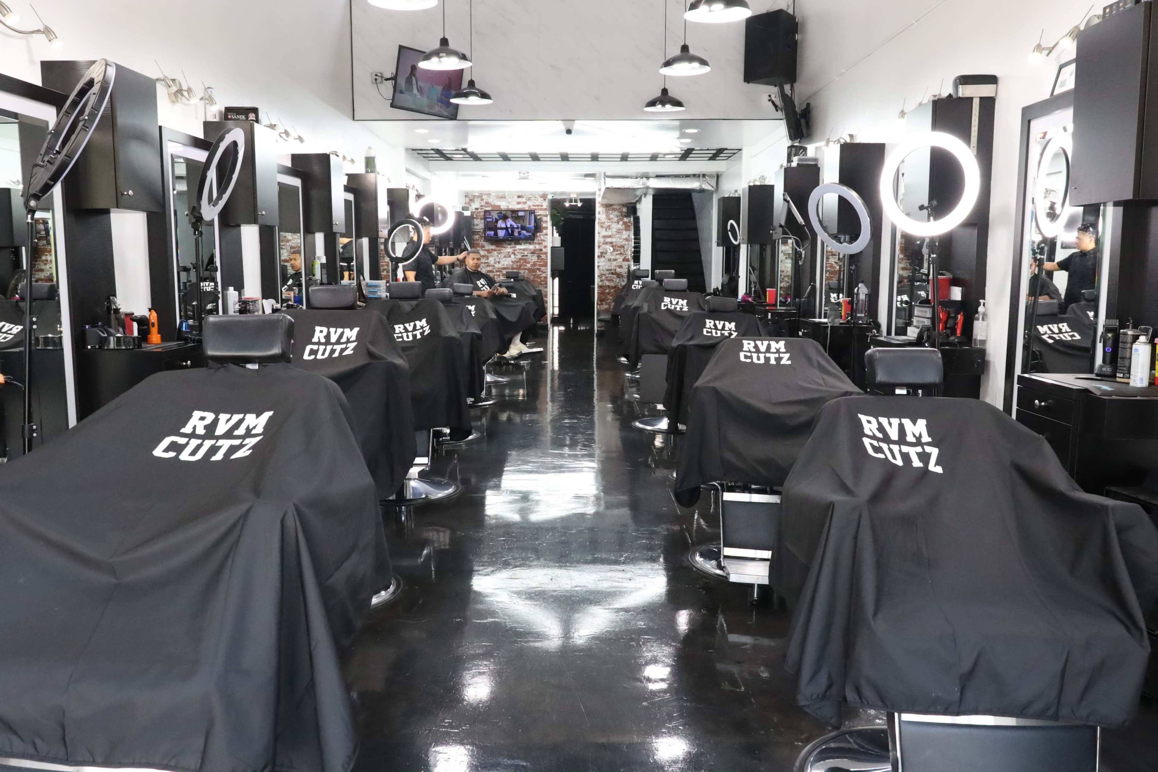 A barbershop interior with multiple styling stations, each covered with black capes labeled "RVM CUTZ," and bright circular mirrors illuminated above each chair.