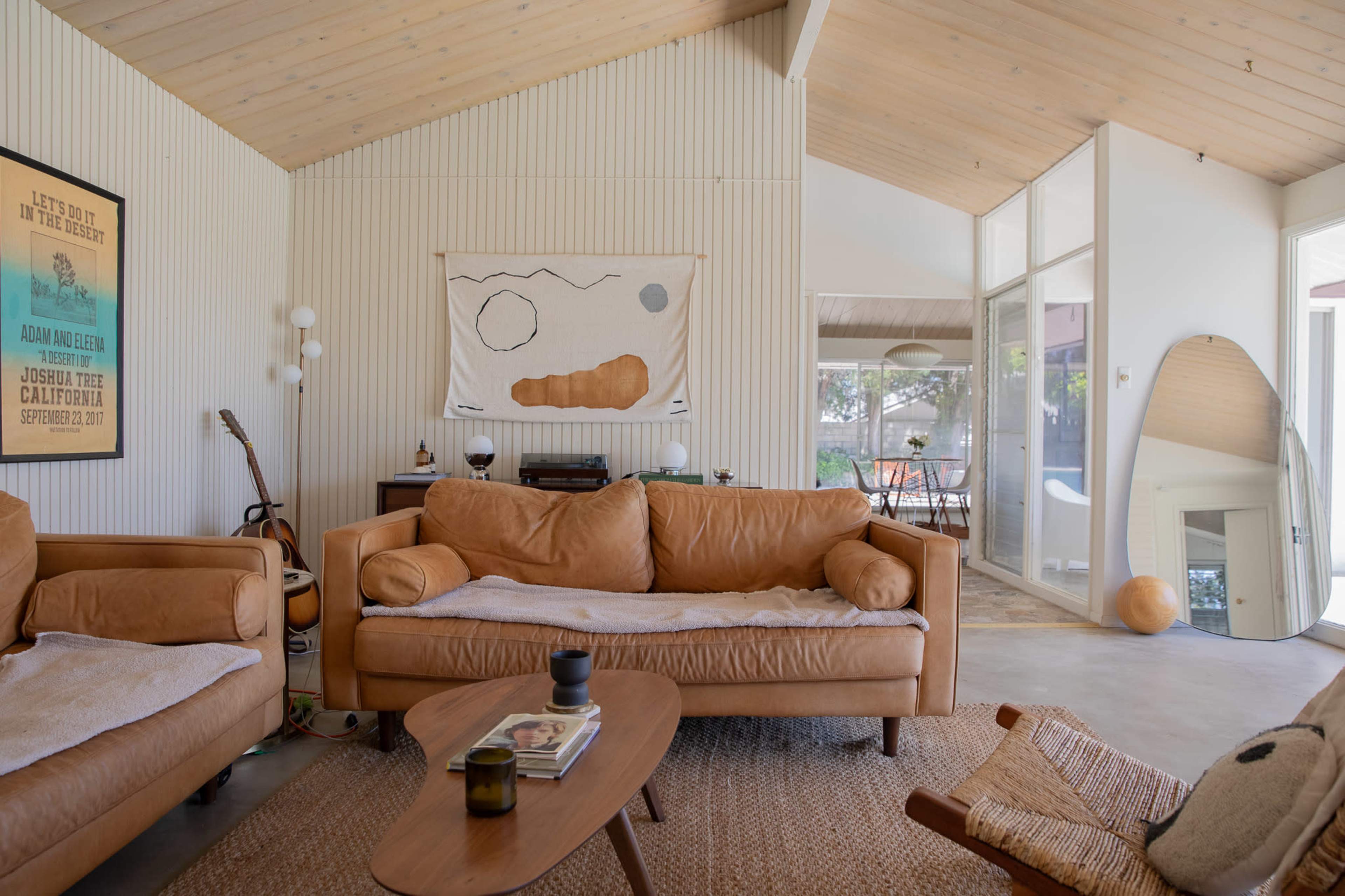 The image shows a cozy living room with leather sofas, a wooden coffee table, and large windows overlooking an outdoor space.