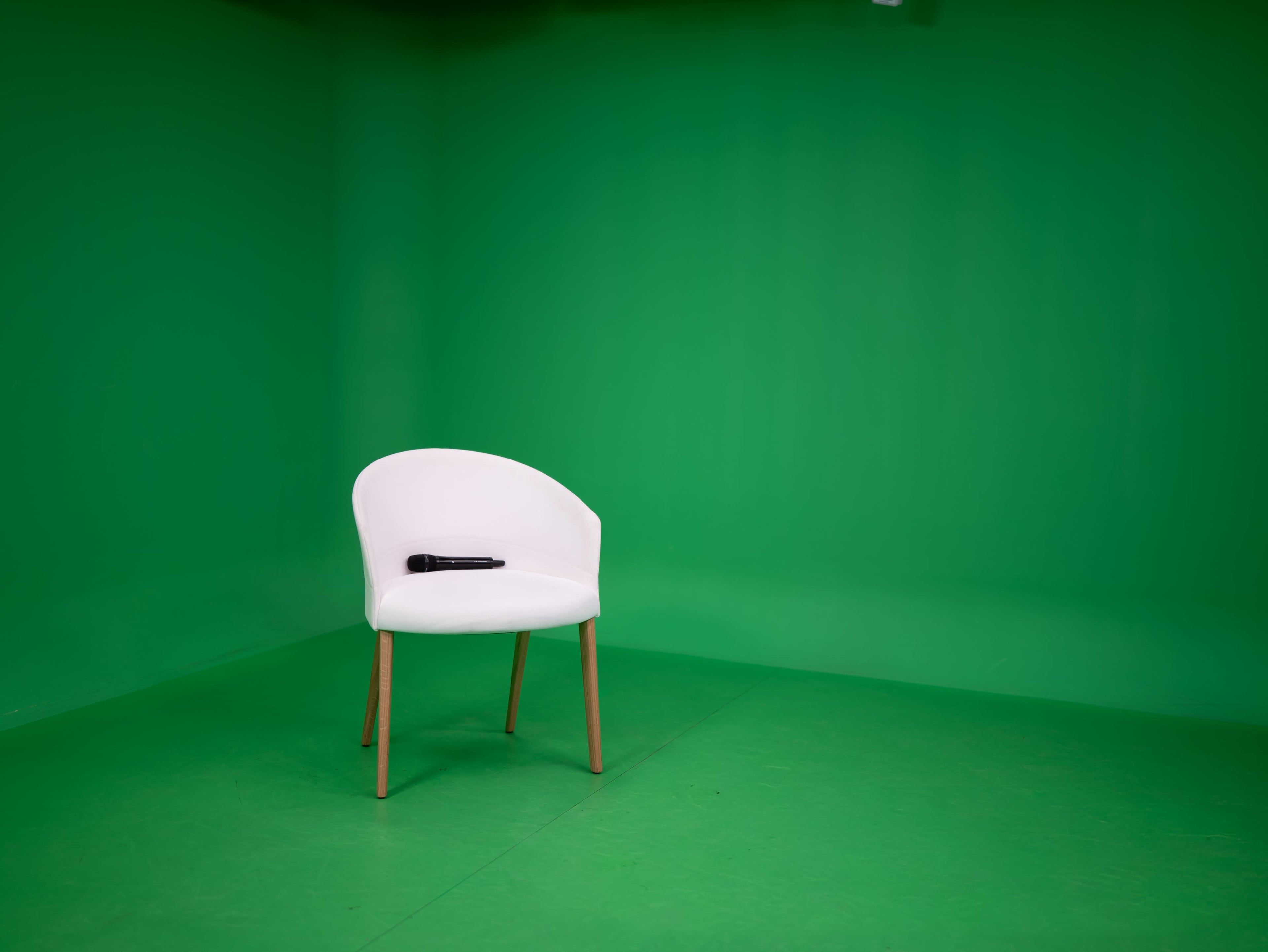 A white chair with wooden legs sits in front of a green backdrop, with a black microphone placed on it.