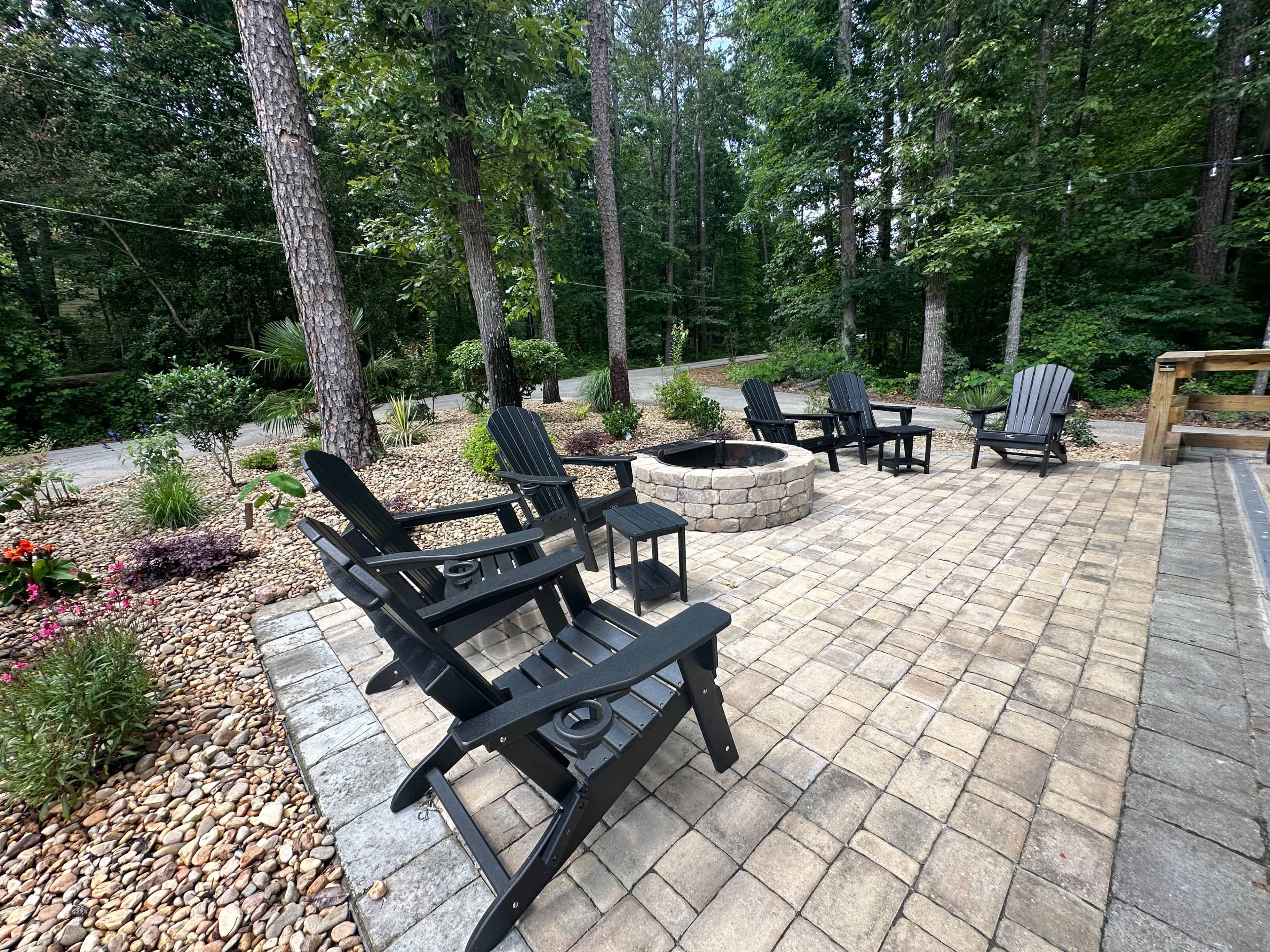 A stone patio area surrounded by trees, featuring several black Adirondack chairs arranged around a fire pit.