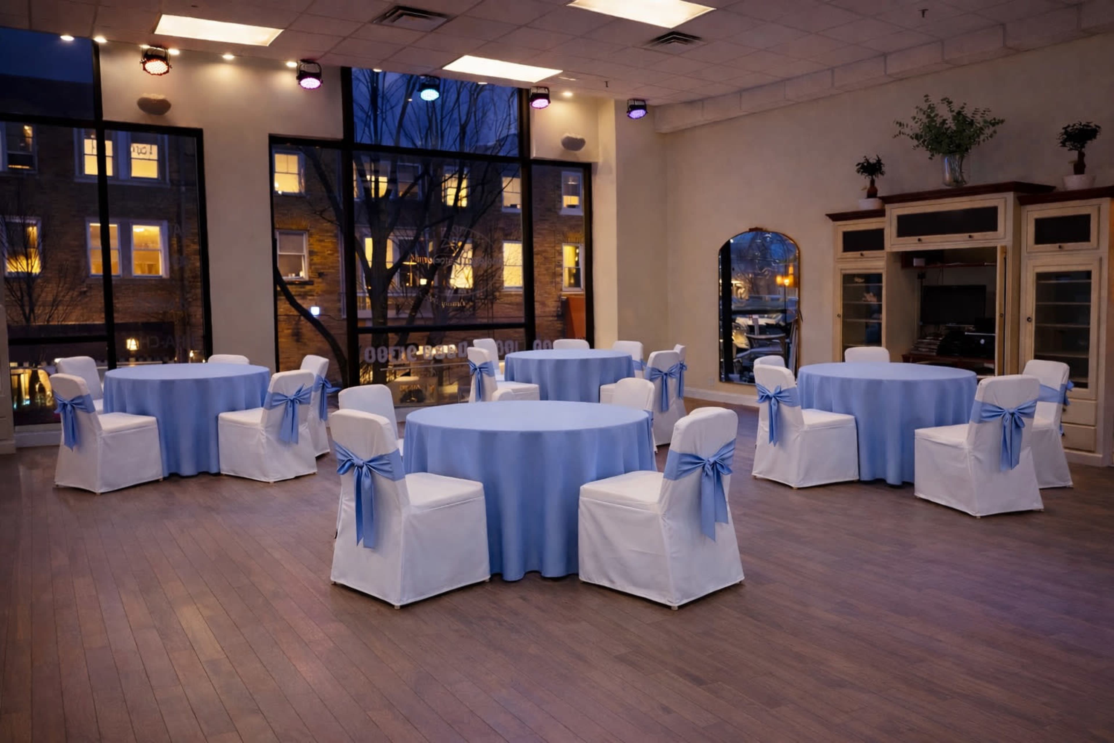 The image shows a banquet hall with round tables covered in light blue tablecloths and white chairs adorned with blue bows, set against large windows at dusk.