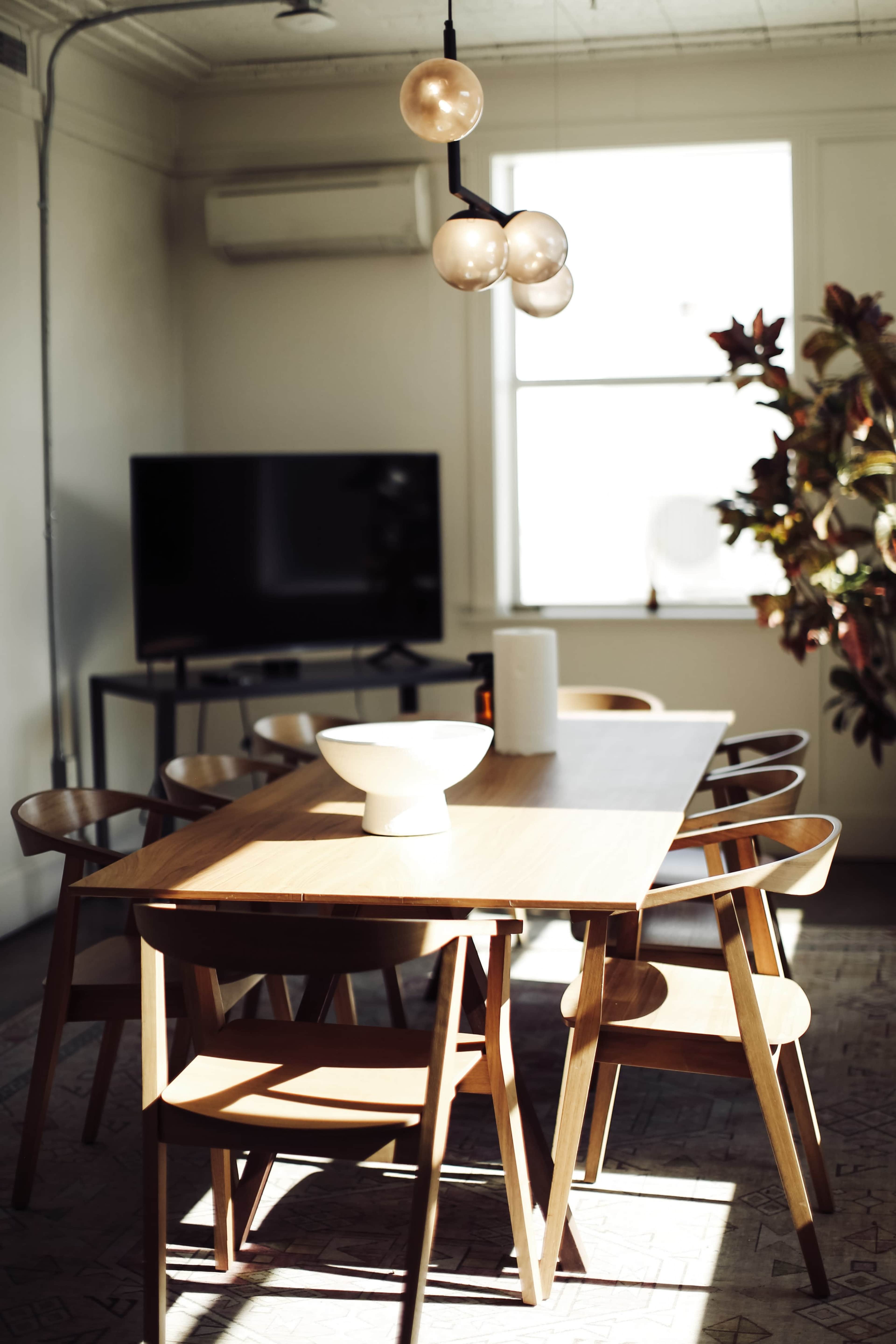 A wooden dining table surrounded by six chairs is displayed in a well-lit room with a television and a large window.