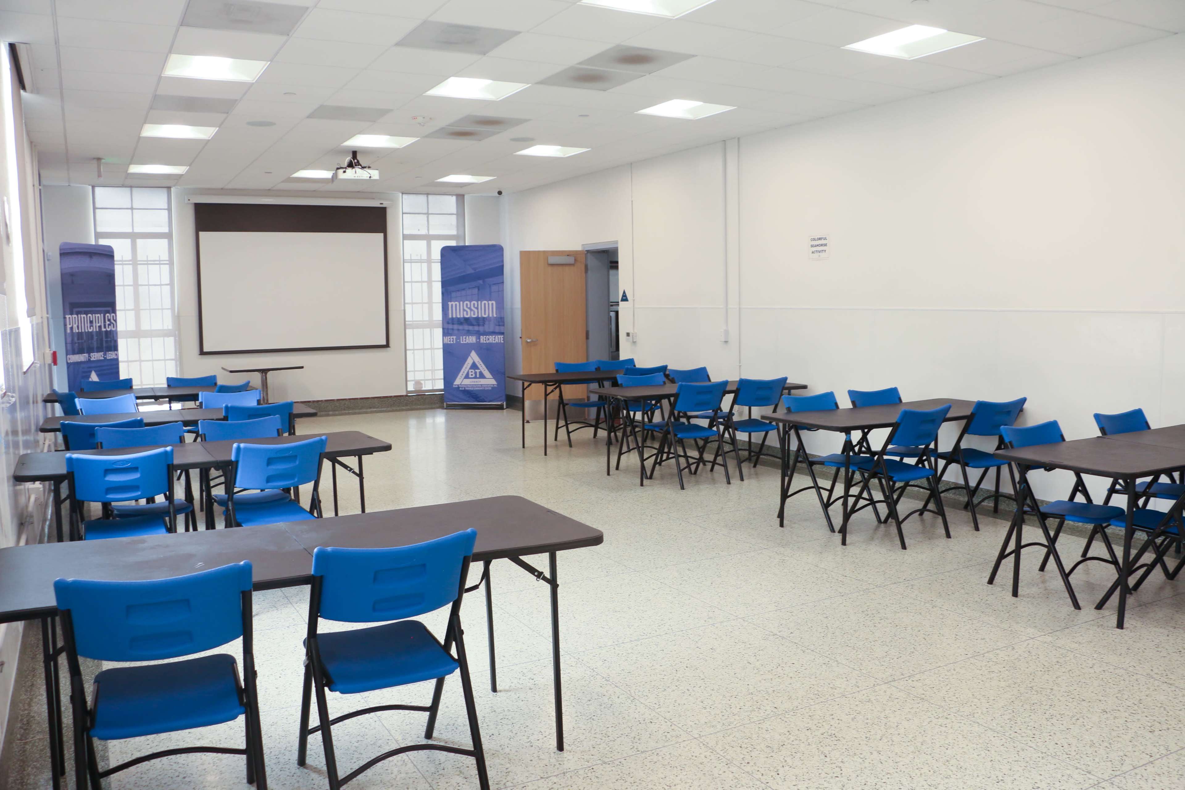The image shows a sparsely furnished classroom with blue chairs and tables arranged around the space, along with a projector screen at the front.
