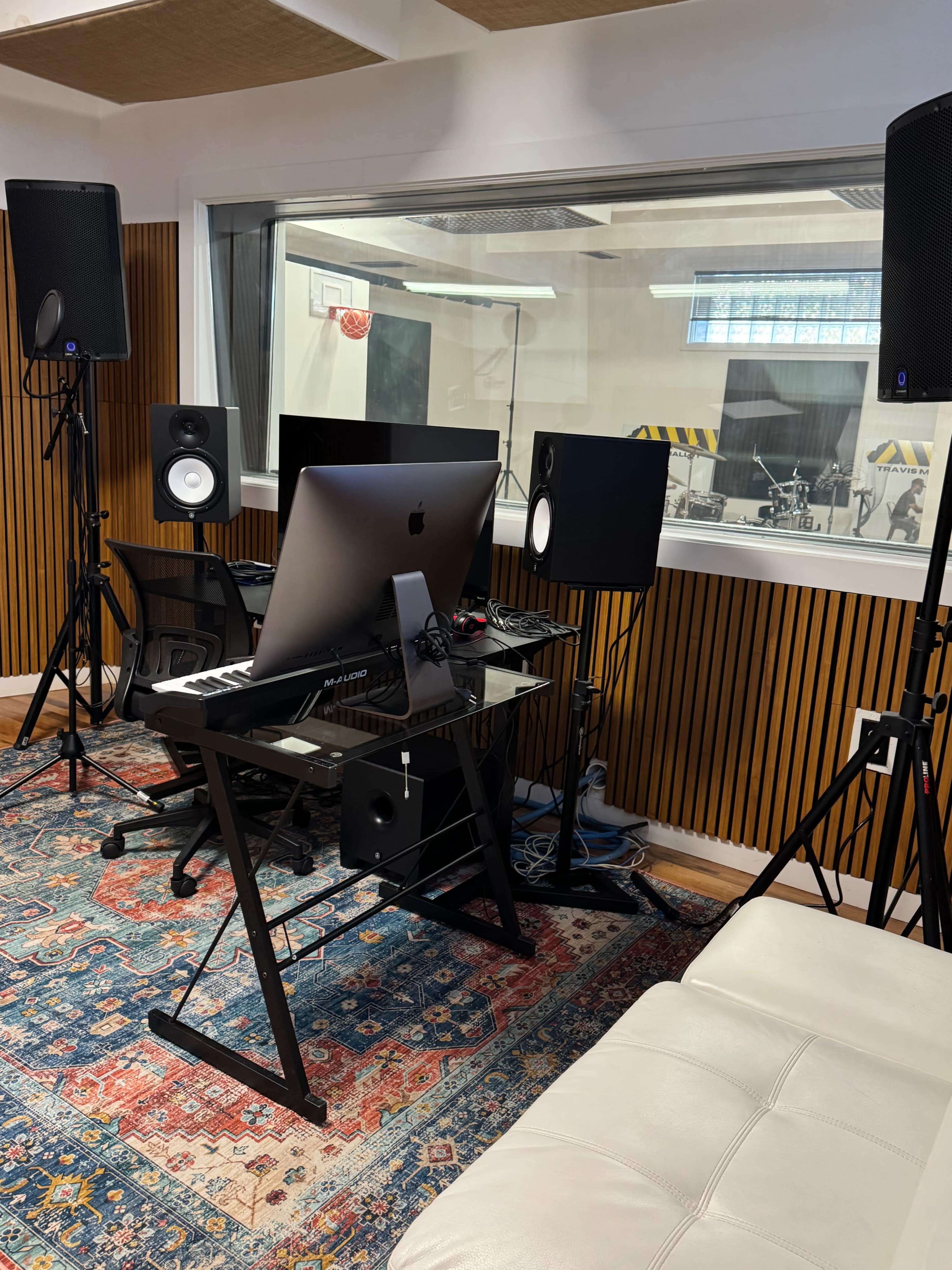 The image shows a modern music production studio with a computer setup, studio monitors, and a visible recording area behind glass.