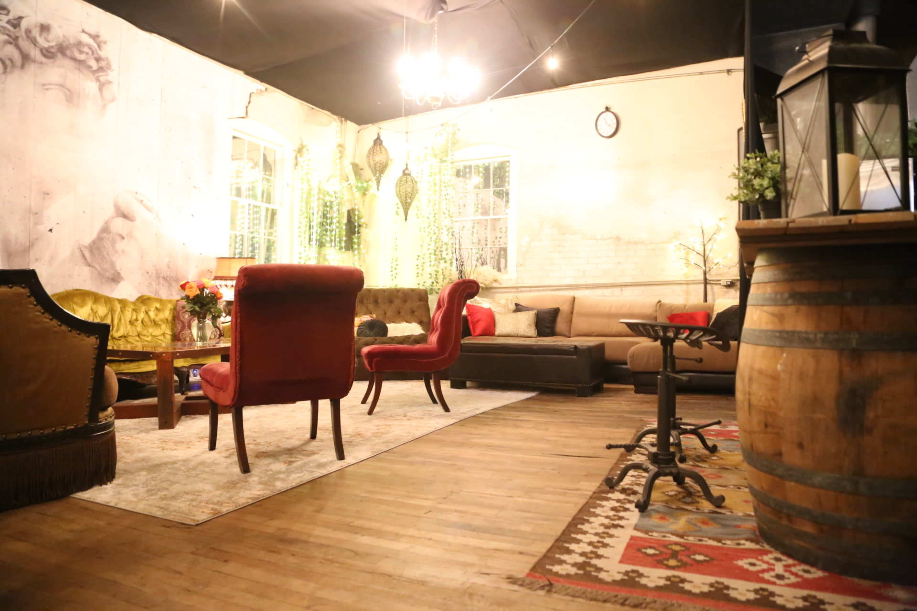 The scene depicts a cozy interior space featuring vintage furniture, including plush red chairs, a yellow sofa, and a wooden barrel, illuminated by soft lighting.