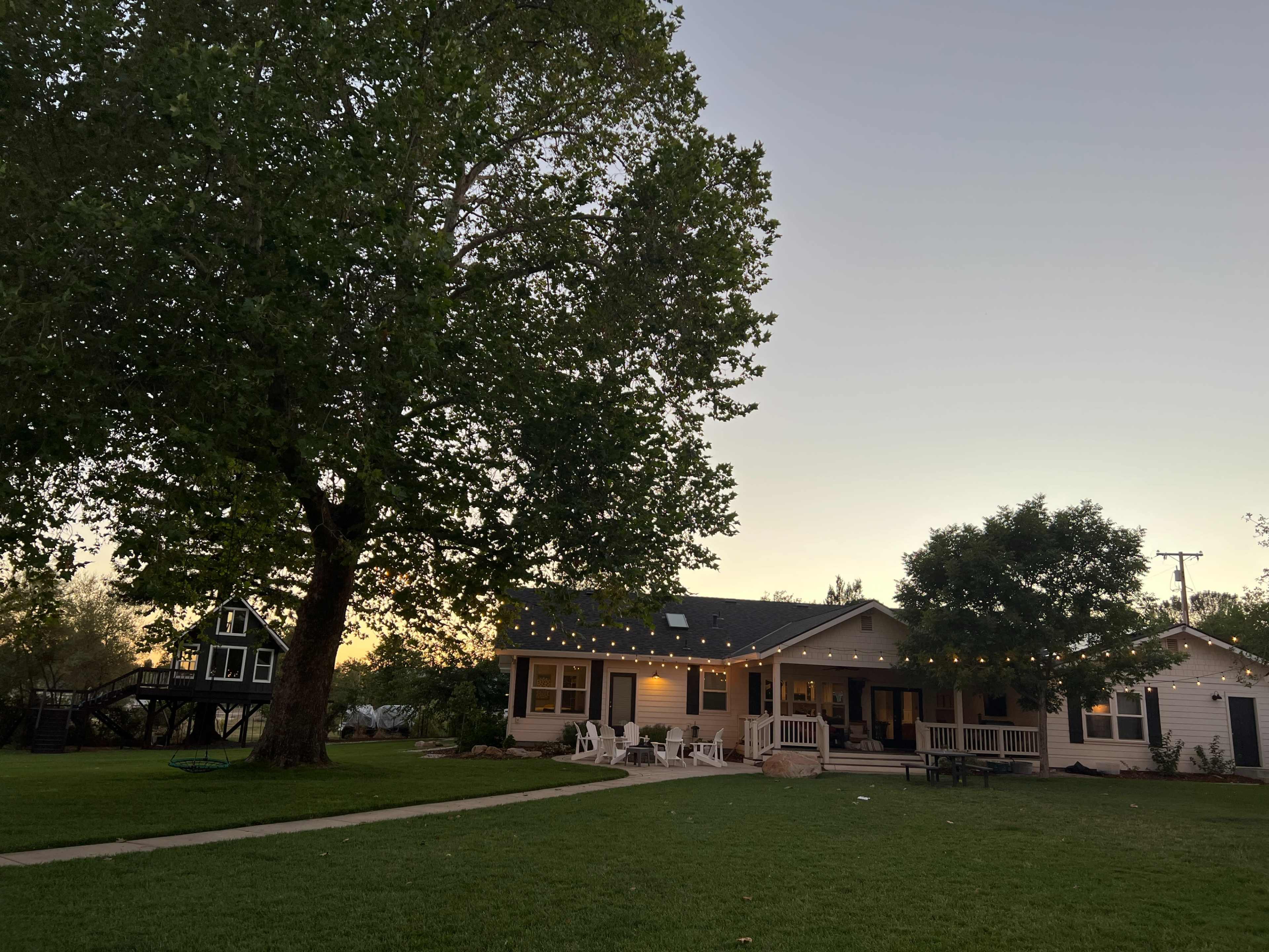 A large house with a porch is illuminated by string lights, surrounded by trees and a lawn at dusk.