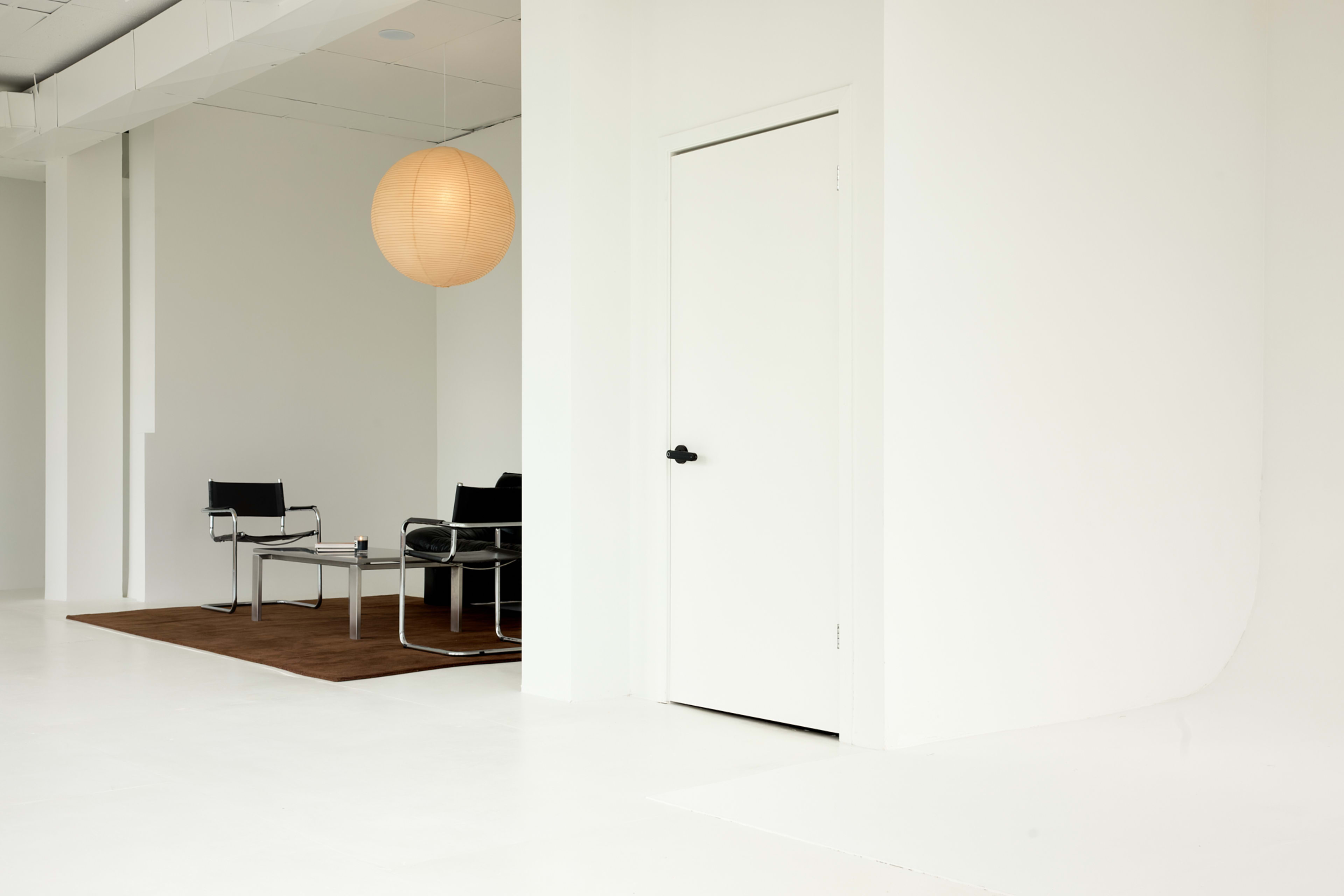 The image shows a minimalist interior space with a white wall, a closed door, a round light fixture, and a black chair set on a brown rug.