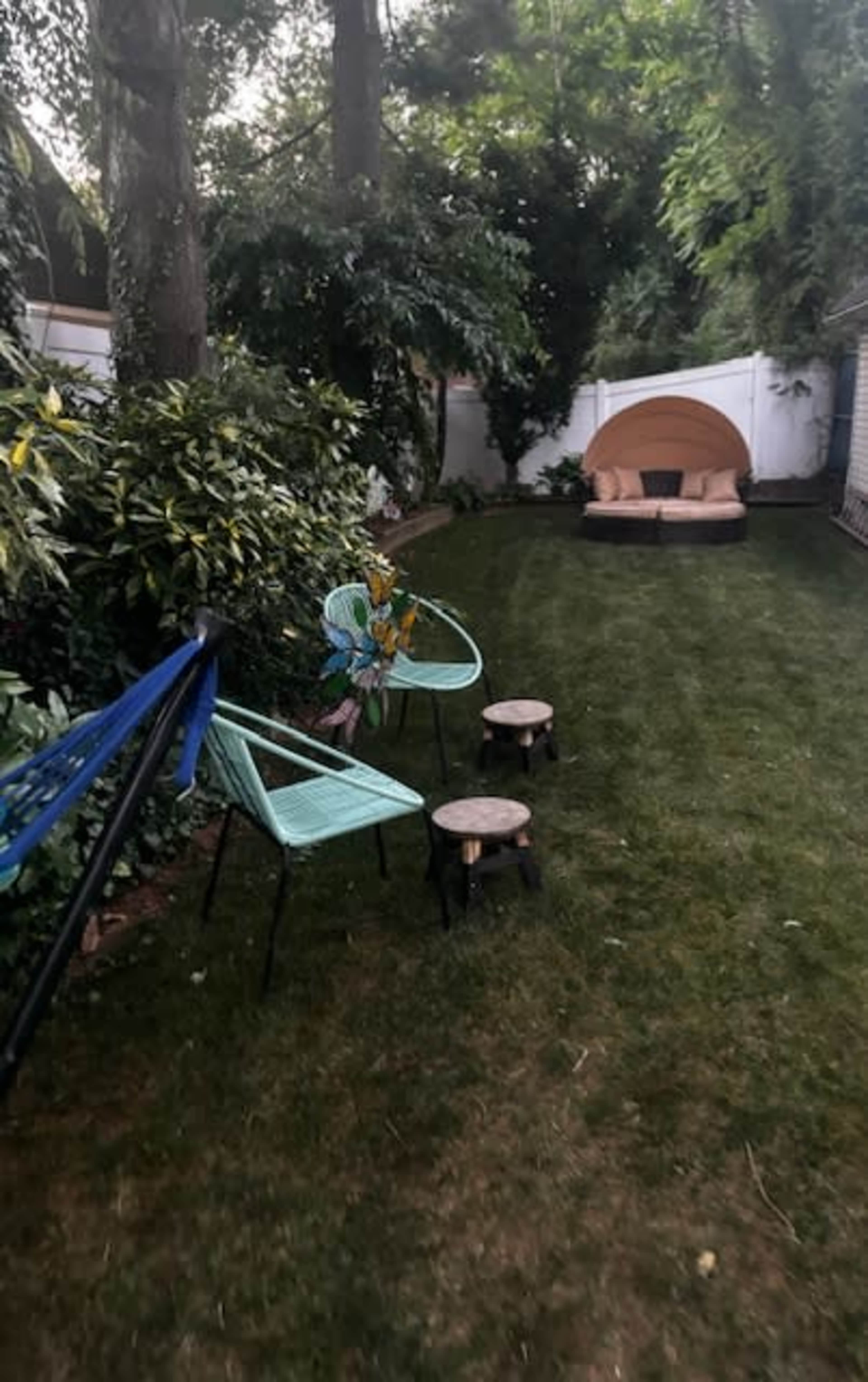 A green backyard features two teal chairs, small wooden stools, a hammock, and a cozy seating area with a canopy.