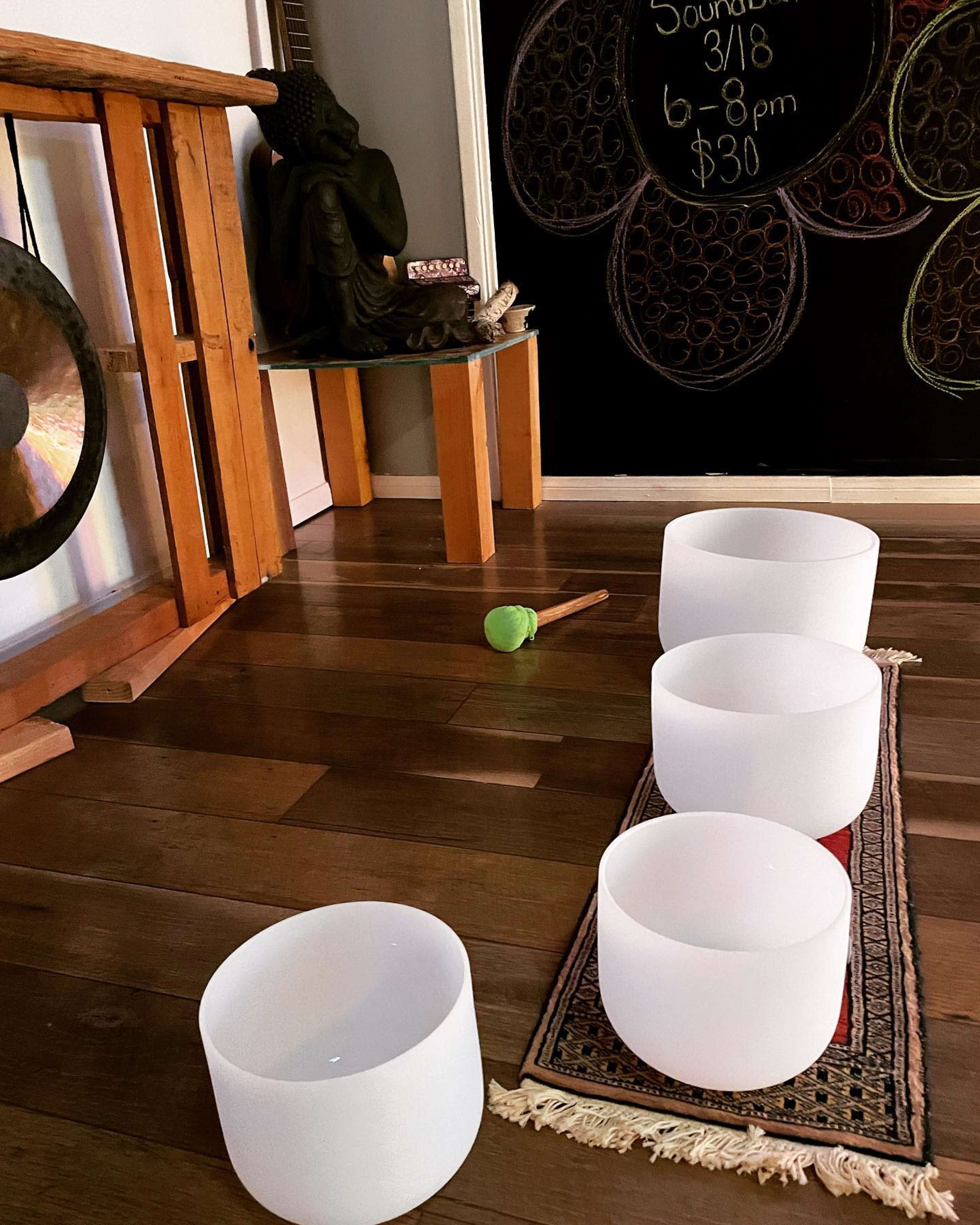 Four frosted crystal singing bowls arranged on a rug in a room that features a wooden structure, a statue, and a chalkboard with event details.