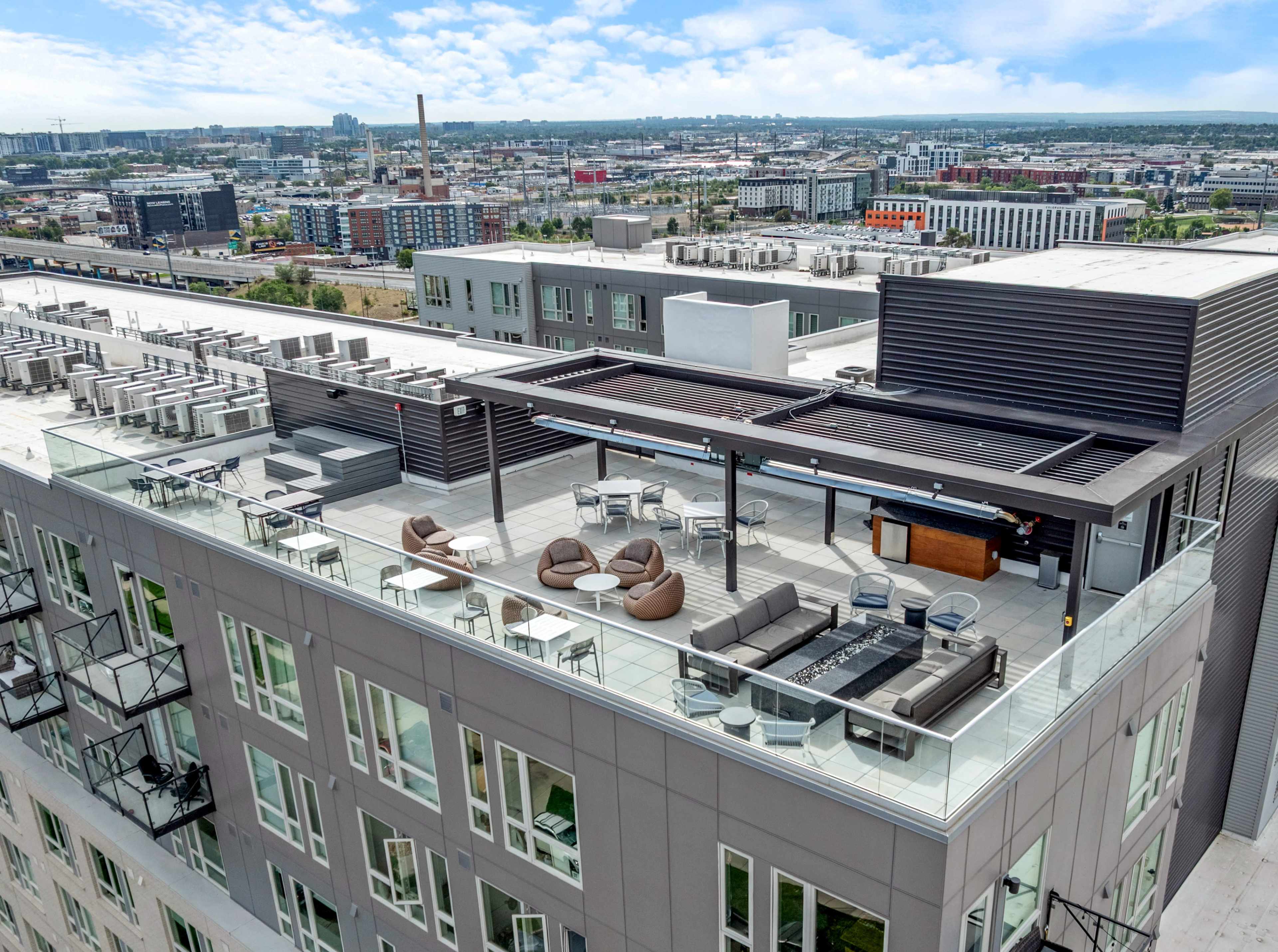 The image shows a modern rooftop terrace with seating areas and a bar overlooking an urban skyline.