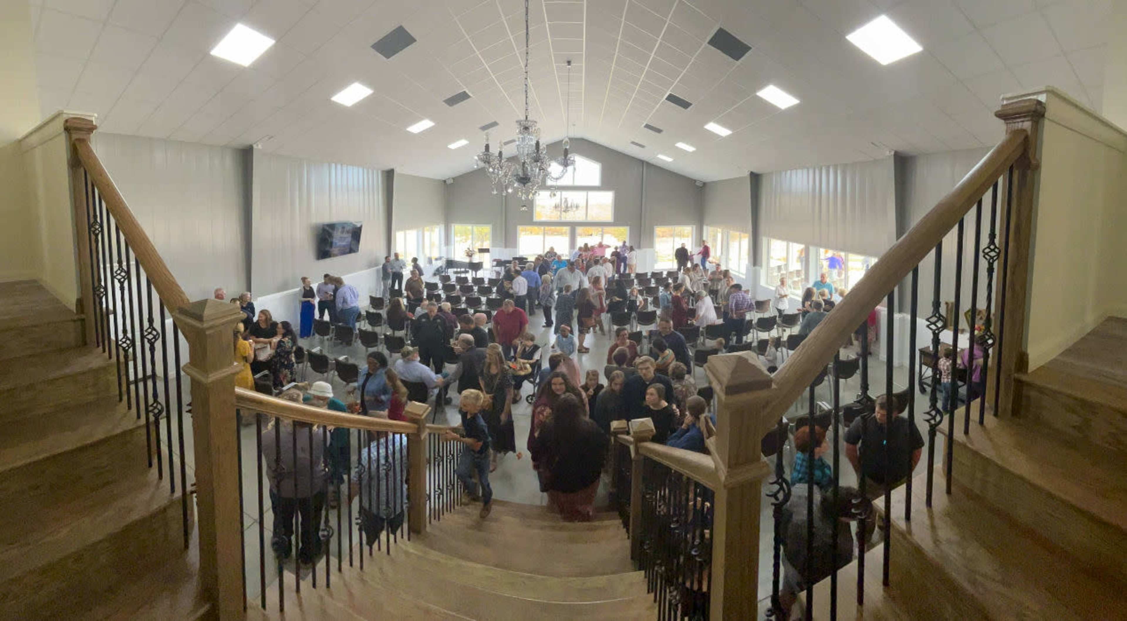 A large group of people gathers in a spacious hall with rows of chairs and a chandelier overhead.