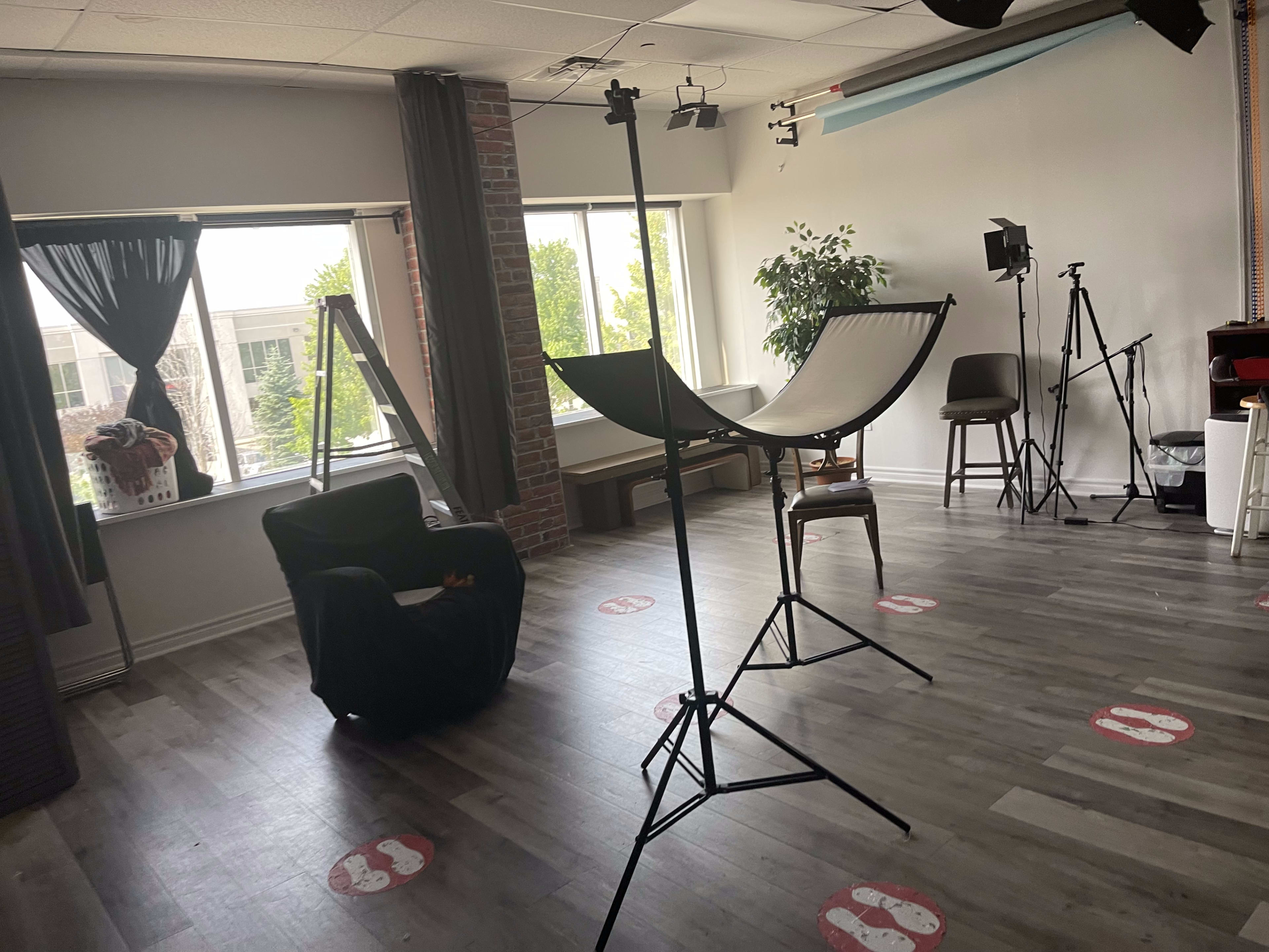A photography studio featuring a black chair, a hammock stand, and several lighting equipment setups, with large windows allowing natural light.