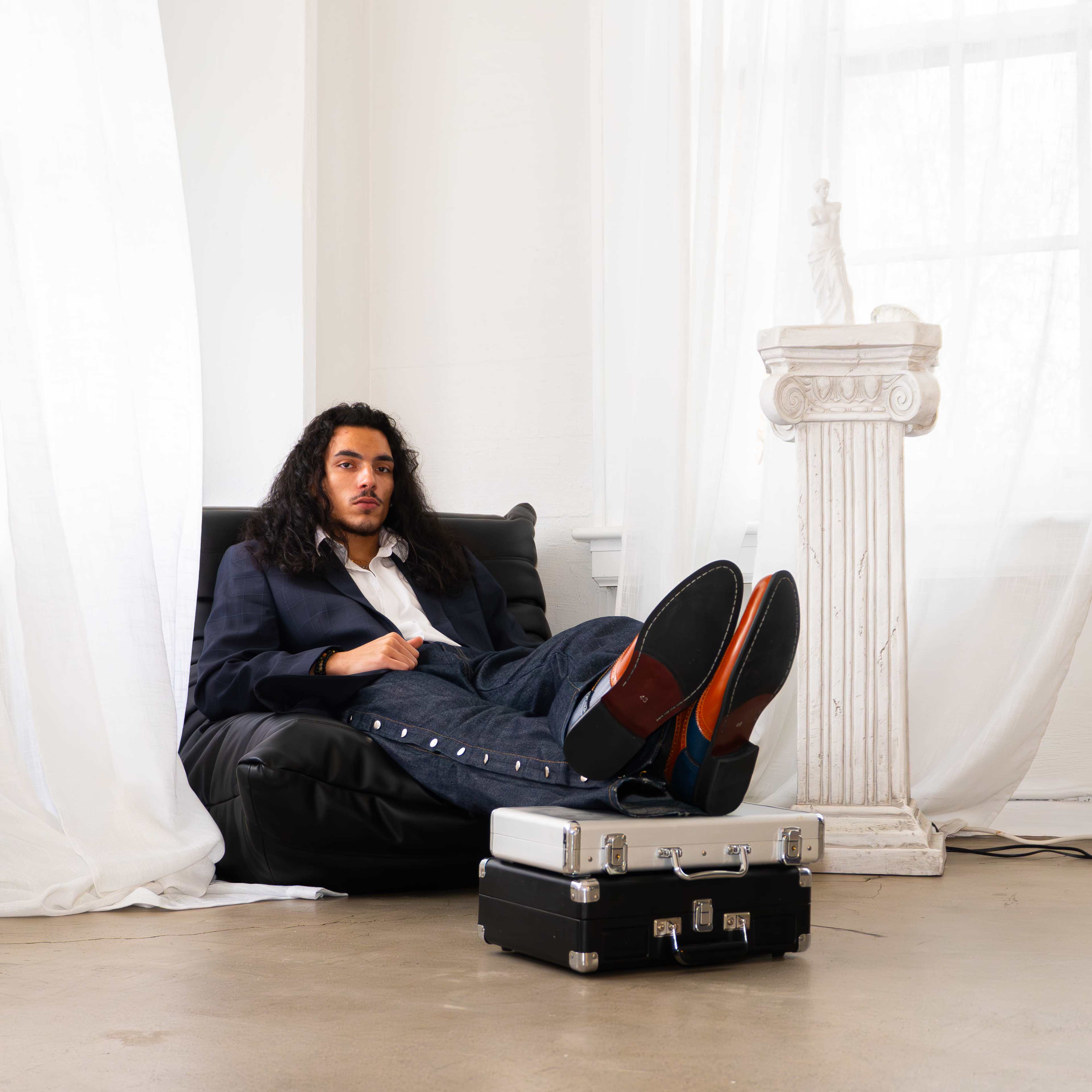 A man with long hair sits casually on a beanbag chair, resting his feet on a black briefcase in a bright, airy room with large windows and sheer curtains.