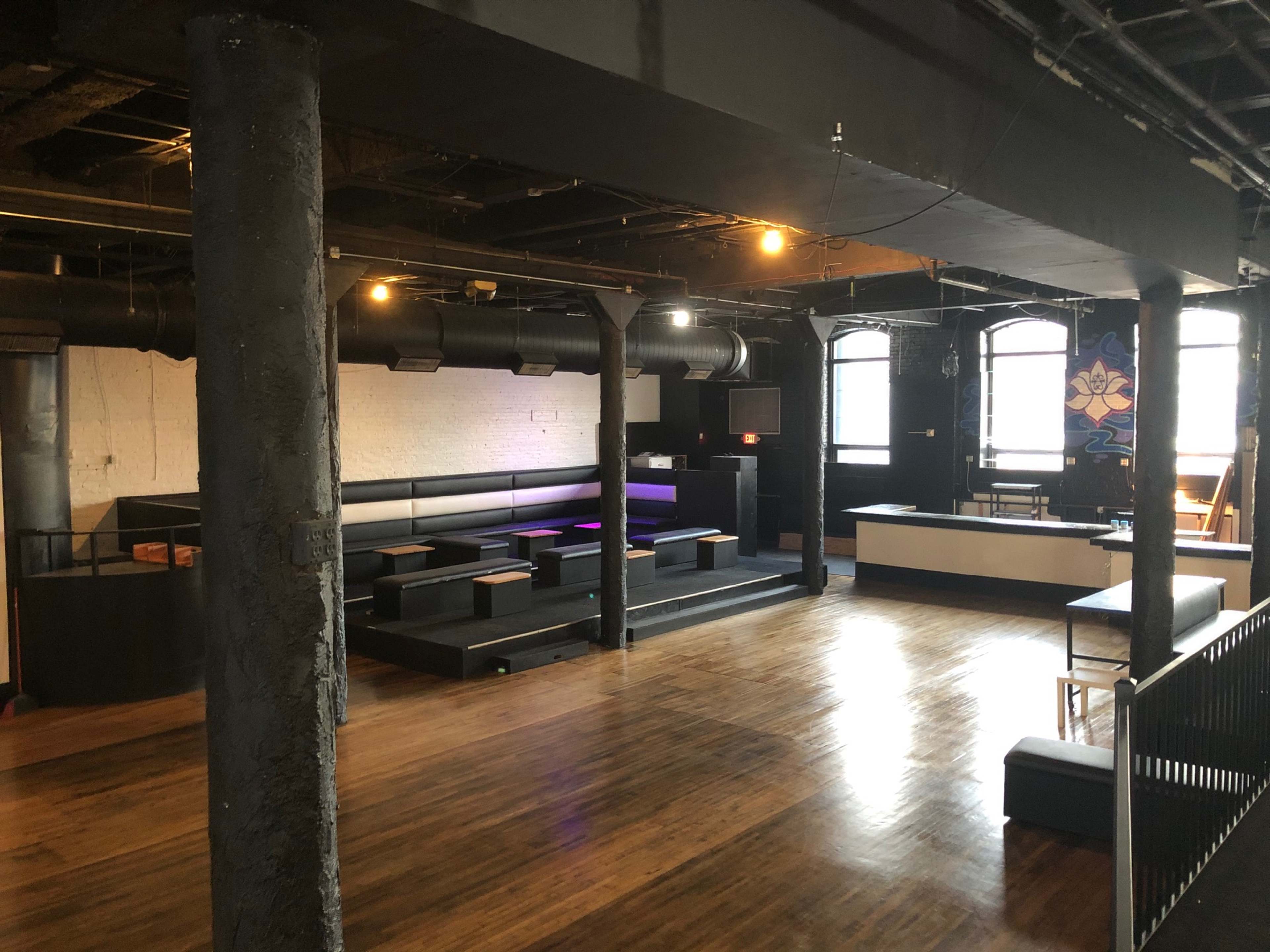 An empty interior space with wooden flooring, black seating along the walls, exposed pipes, and a bar area in the corner.