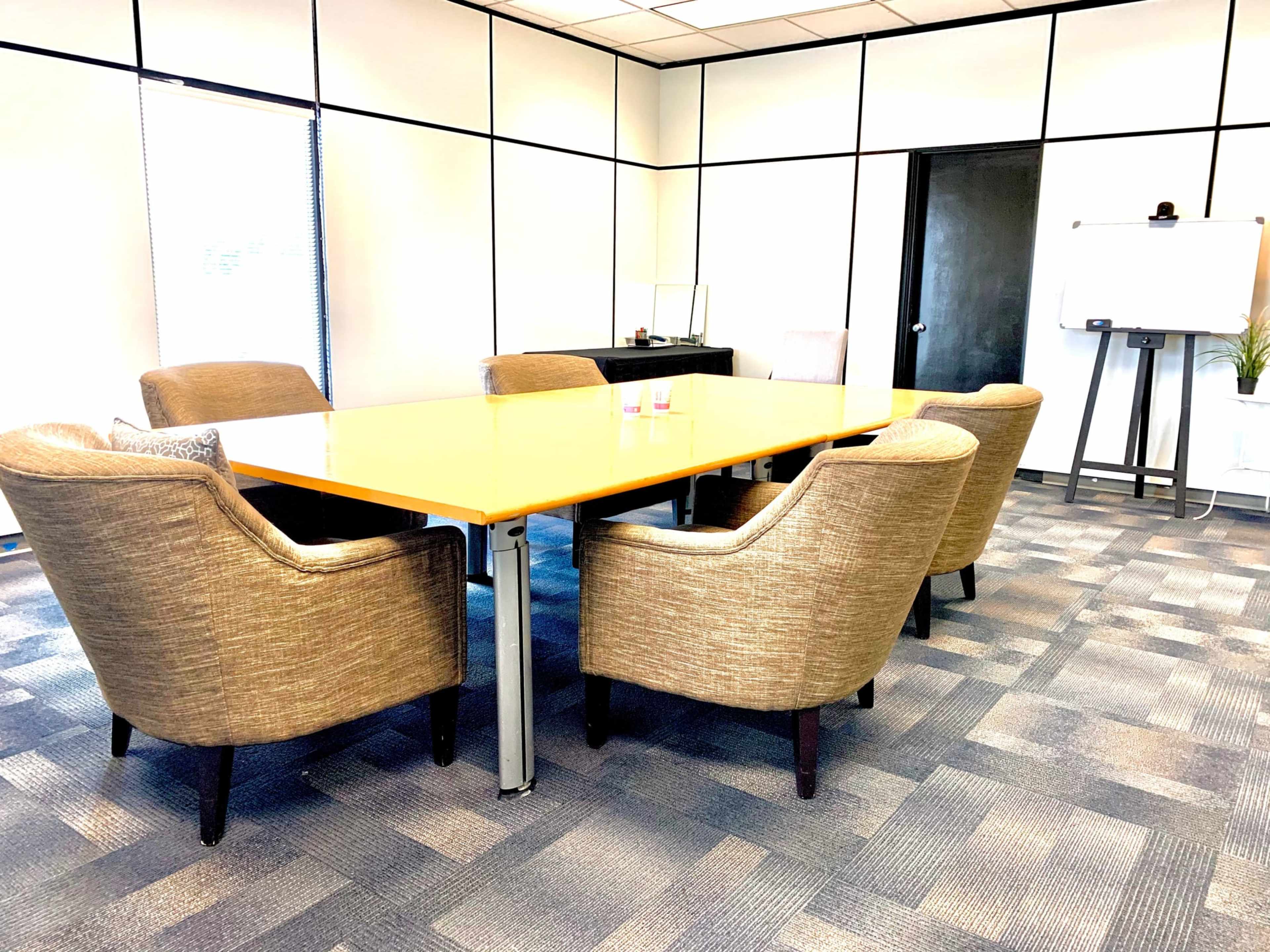 A conference room features a large wooden table surrounded by five upholstered chairs, with a whiteboard and plant in the background.