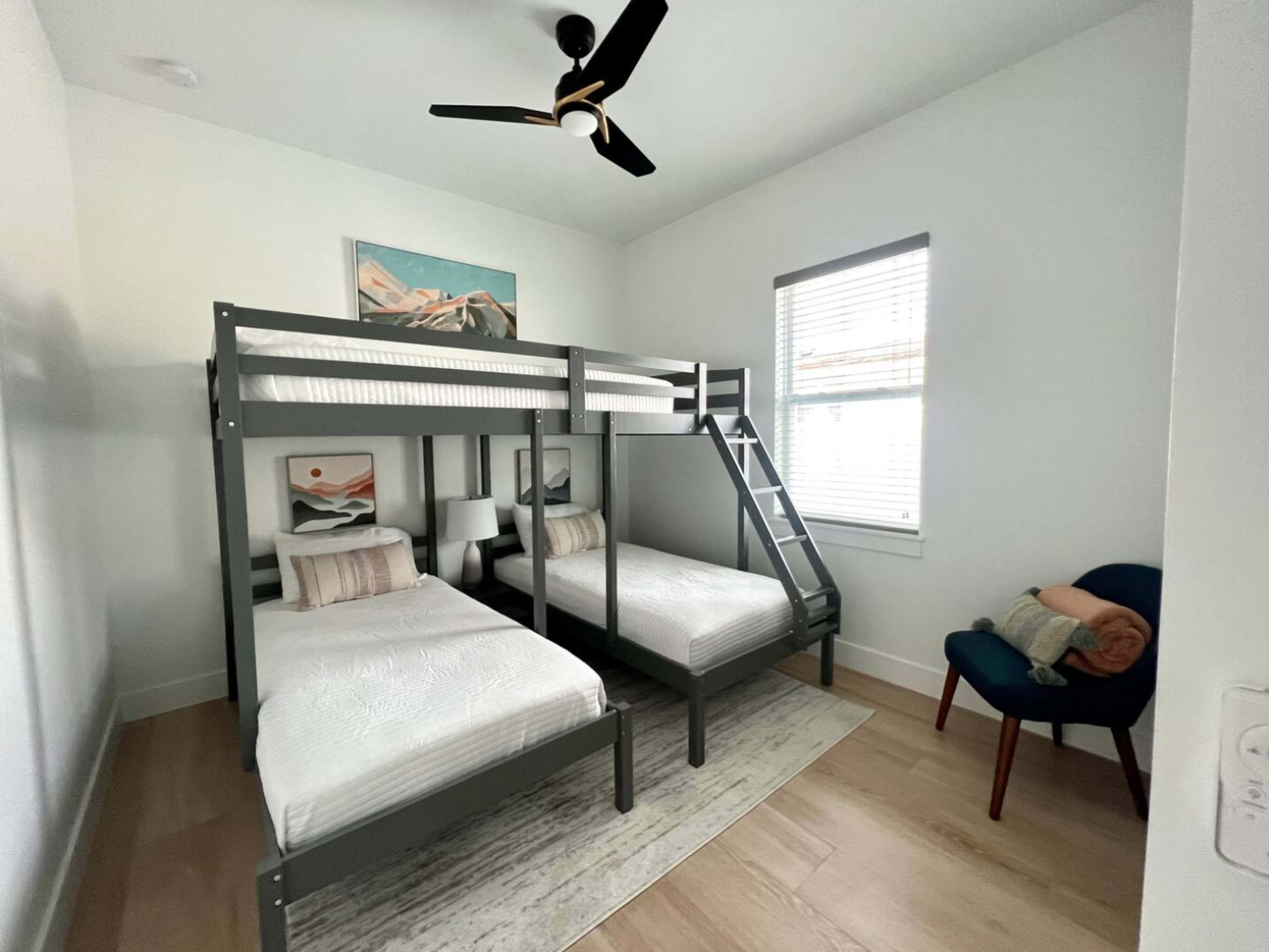 A room features a bunk bed with two twin mattresses and an additional twin bed, accompanied by a chair and a wall-mounted artwork.