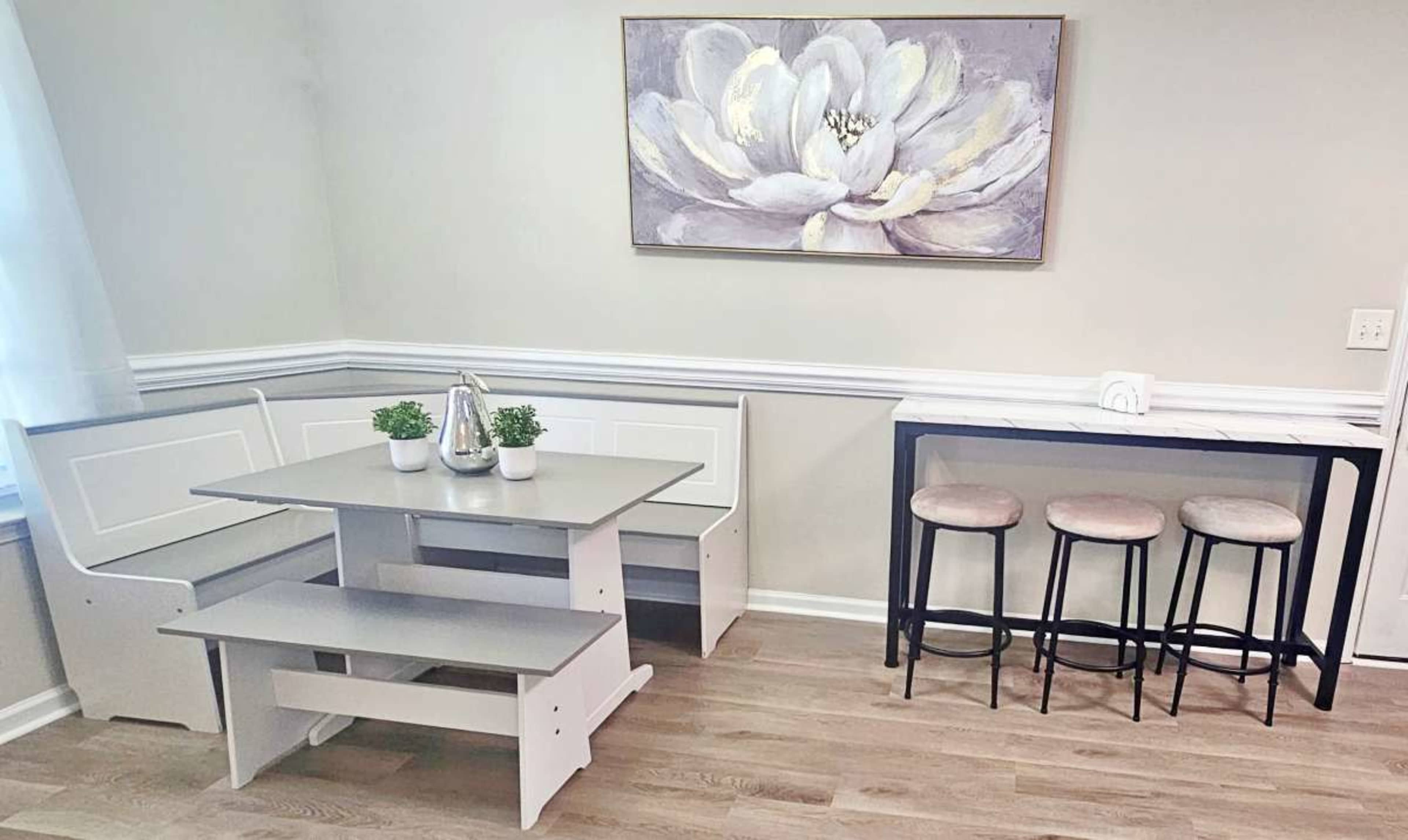 The image shows a dining area featuring a corner bench seating arrangement with a table, two small potted plants on the table, and a separate bar area with three black stools.