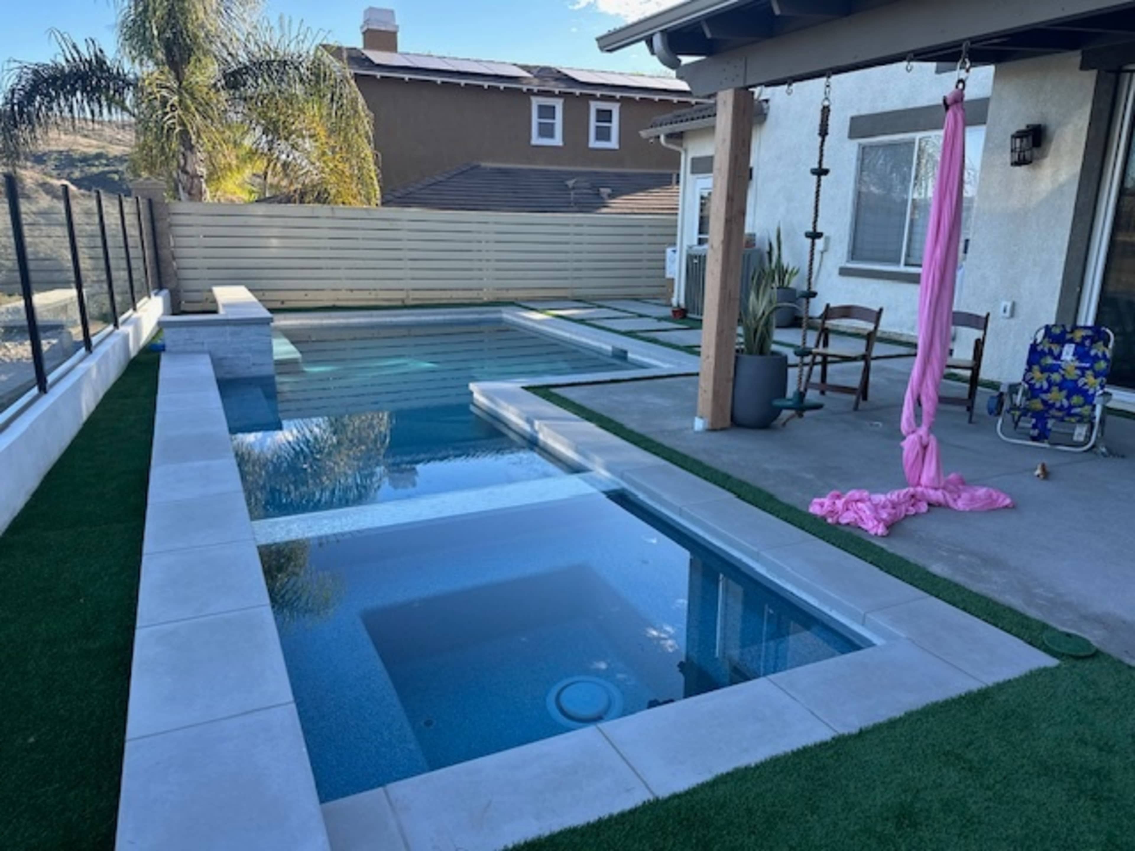 A backyard with a rectangular swimming pool and a spa, surrounded by artificial grass and a patio area with outdoor furniture.