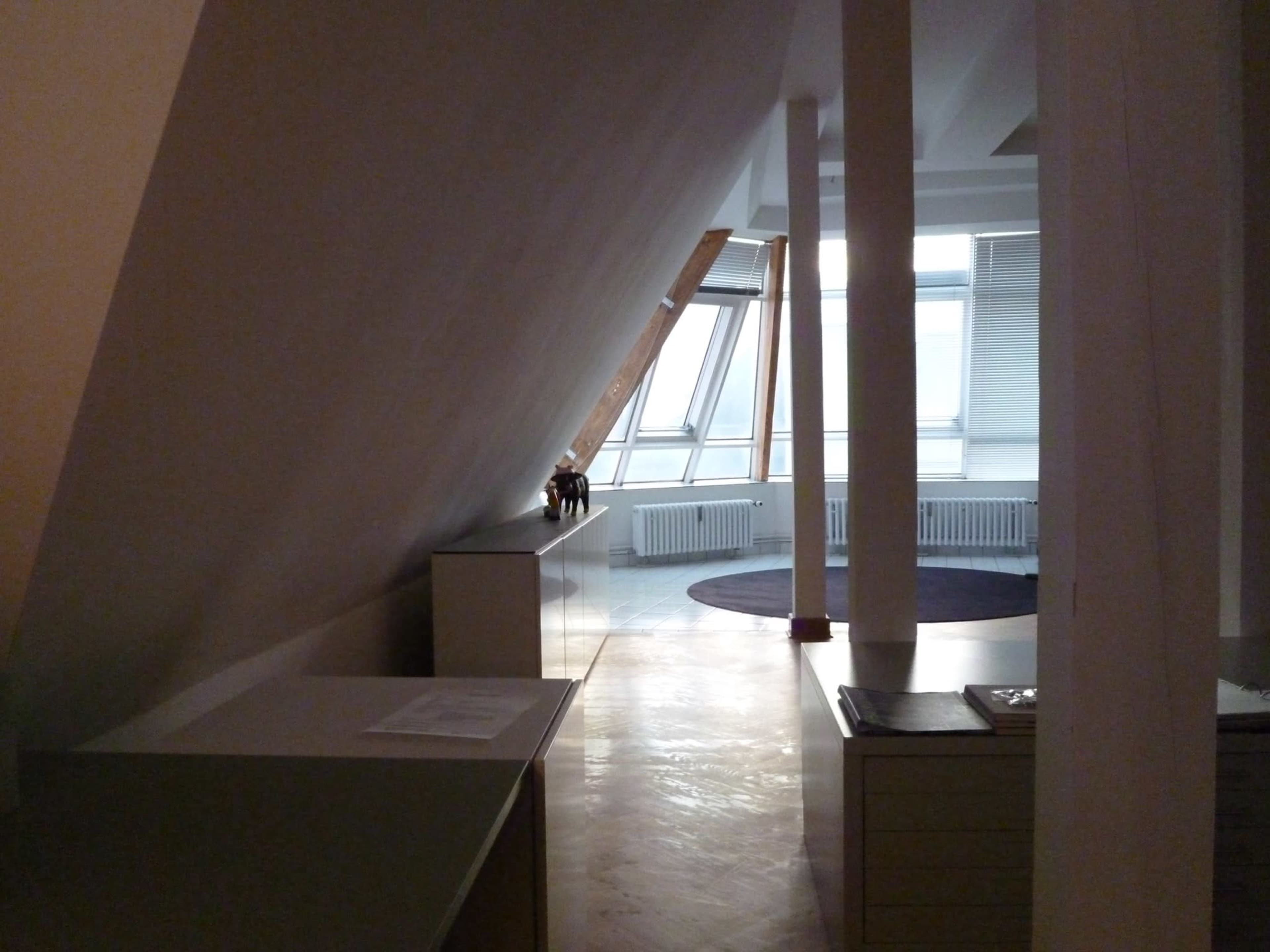 The image shows an interior space with slanted walls, a wooden floor, and furniture along the sides, leading to a large window with a circular rug in the center.