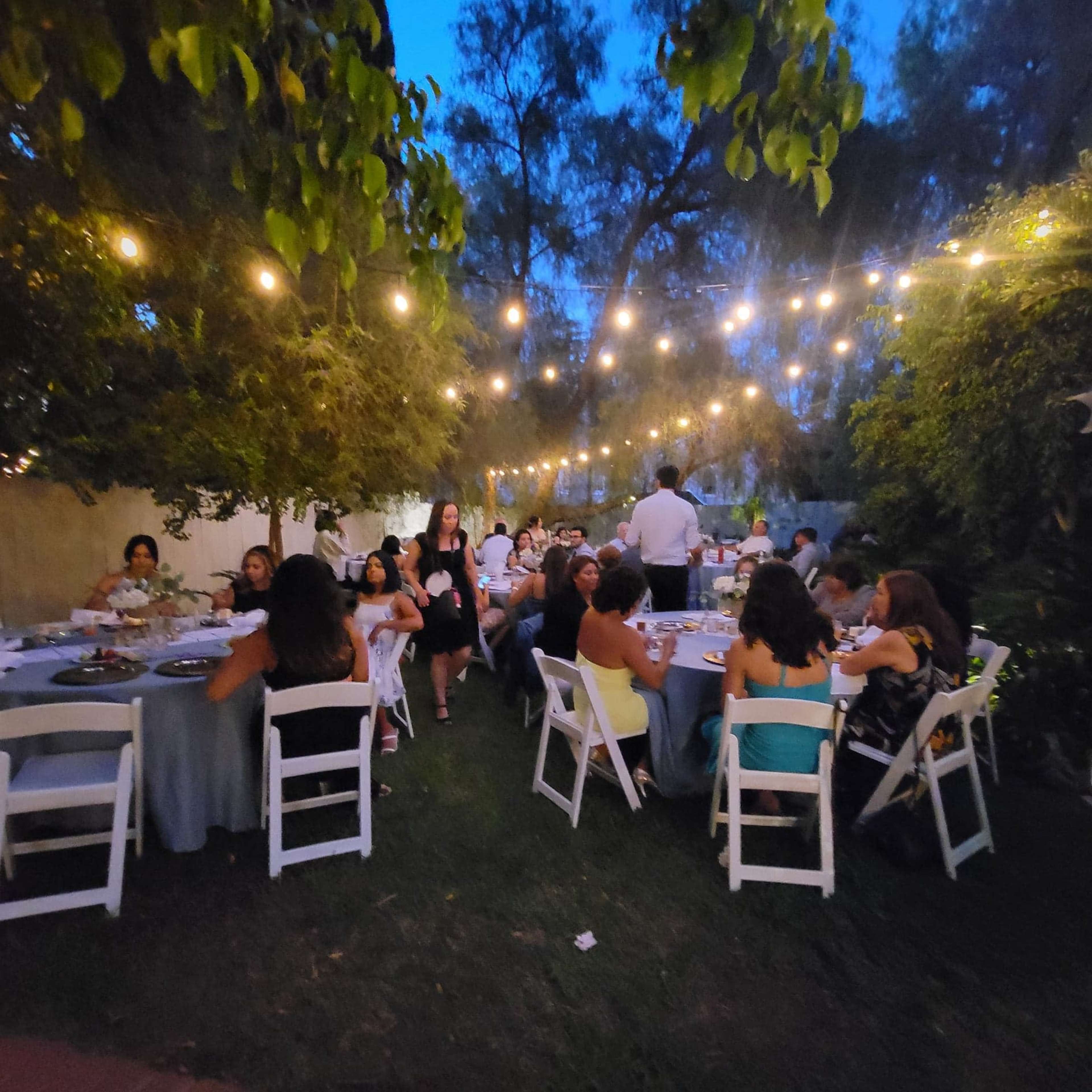 A group of people gathers at tables under string lights in a garden setting during the evening.