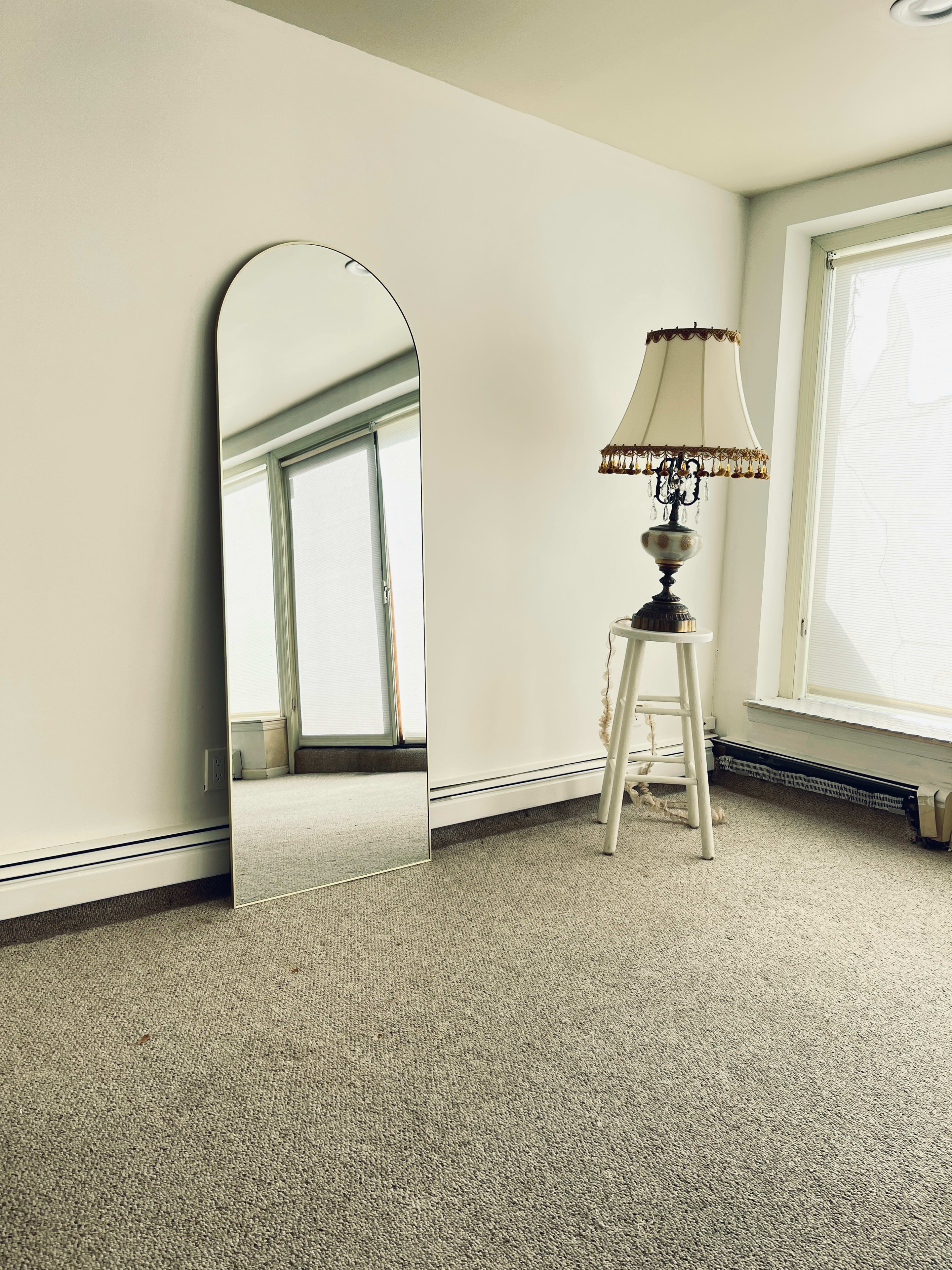 A large arched mirror stands against a wall next to a lamp on a small table in a carpeted, empty room.
