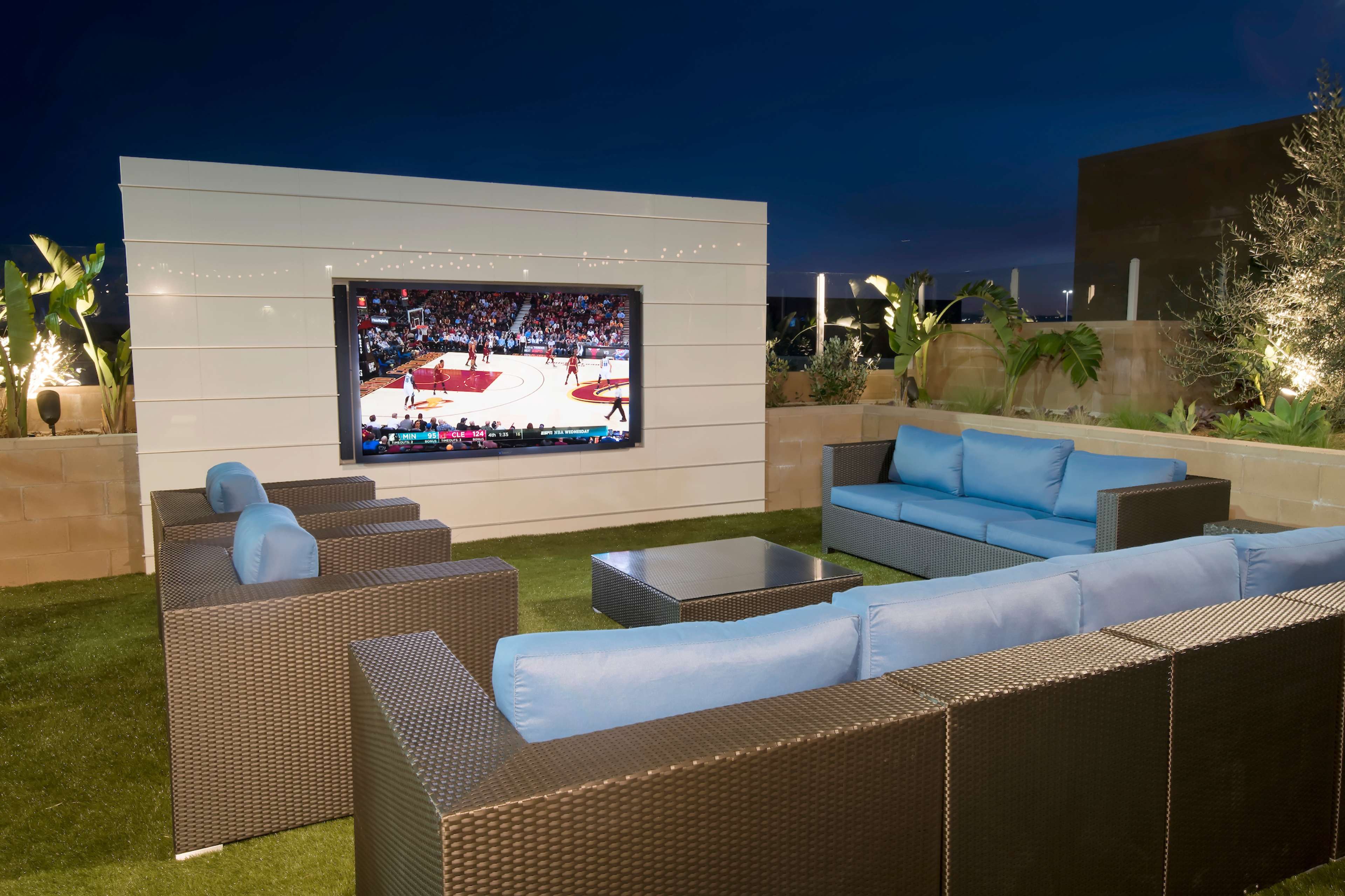 A modern outdoor lounge area features a large screen displaying a basketball game, surrounded by comfortable seating and lush greenery under evening lighting.