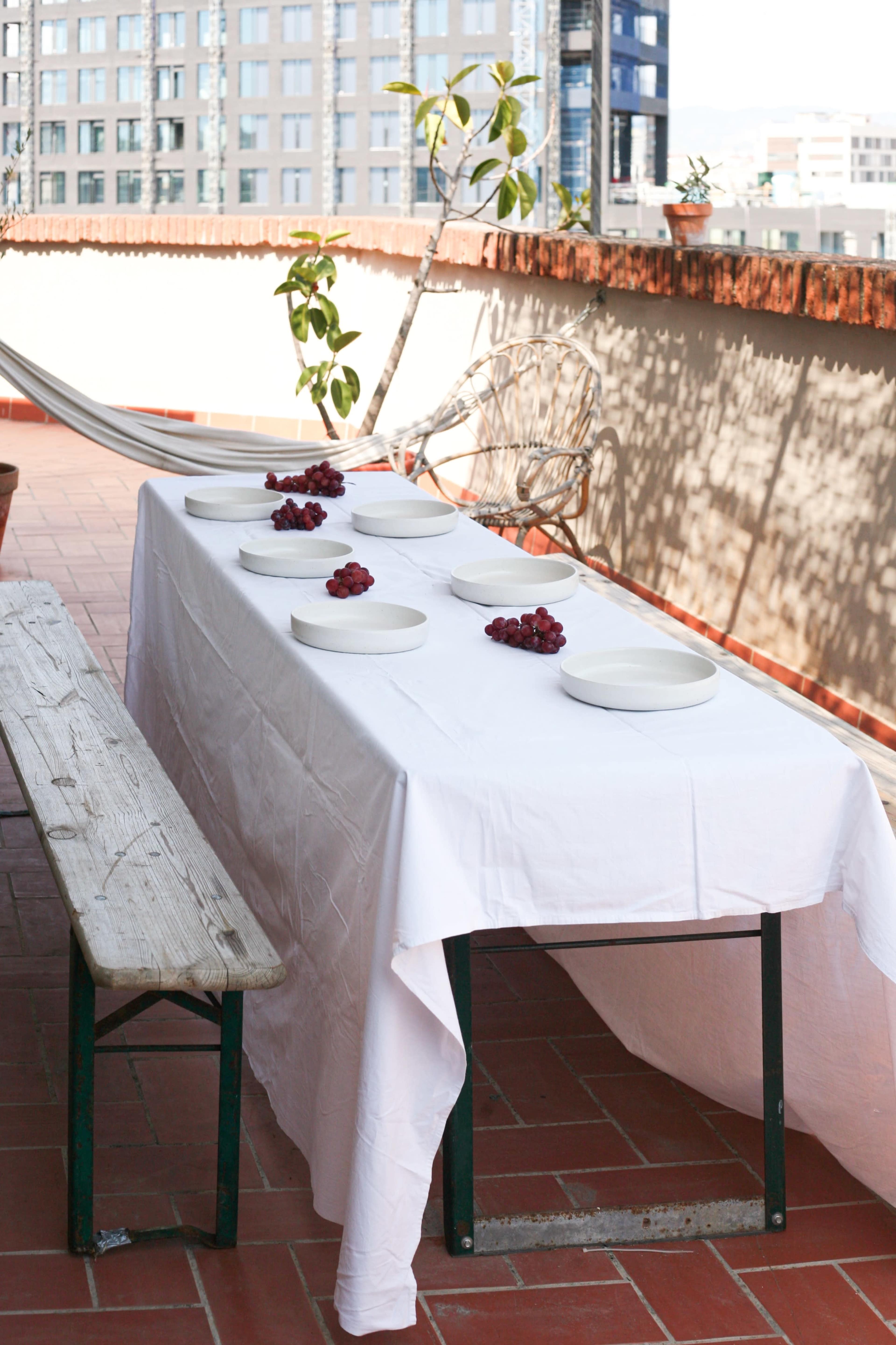 A white tablecloth covers a table set with empty plates and garnished with clusters of grapes on a balcony.