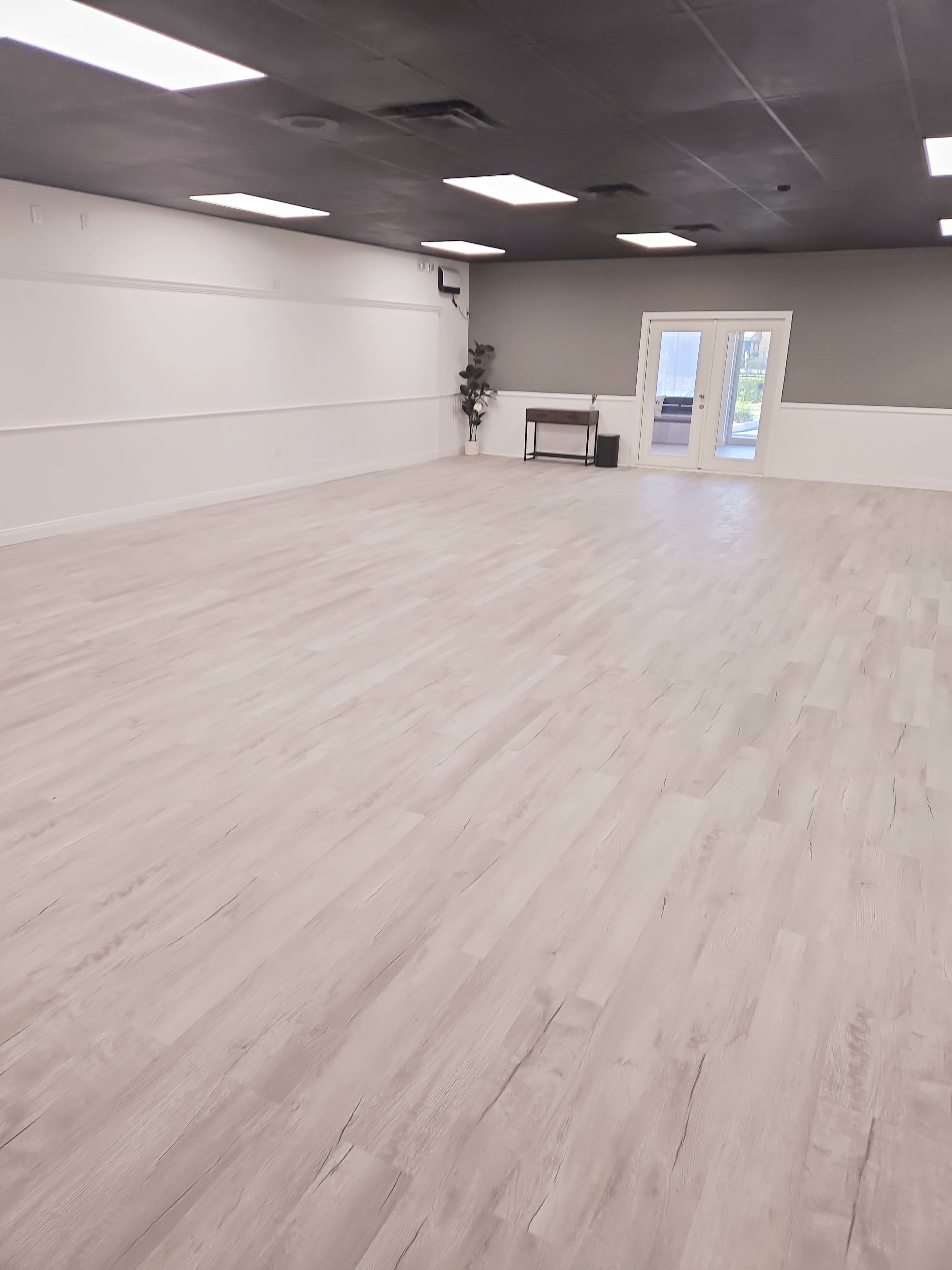 The image shows a spacious, empty room with light-colored wooden flooring and large windows at one end.