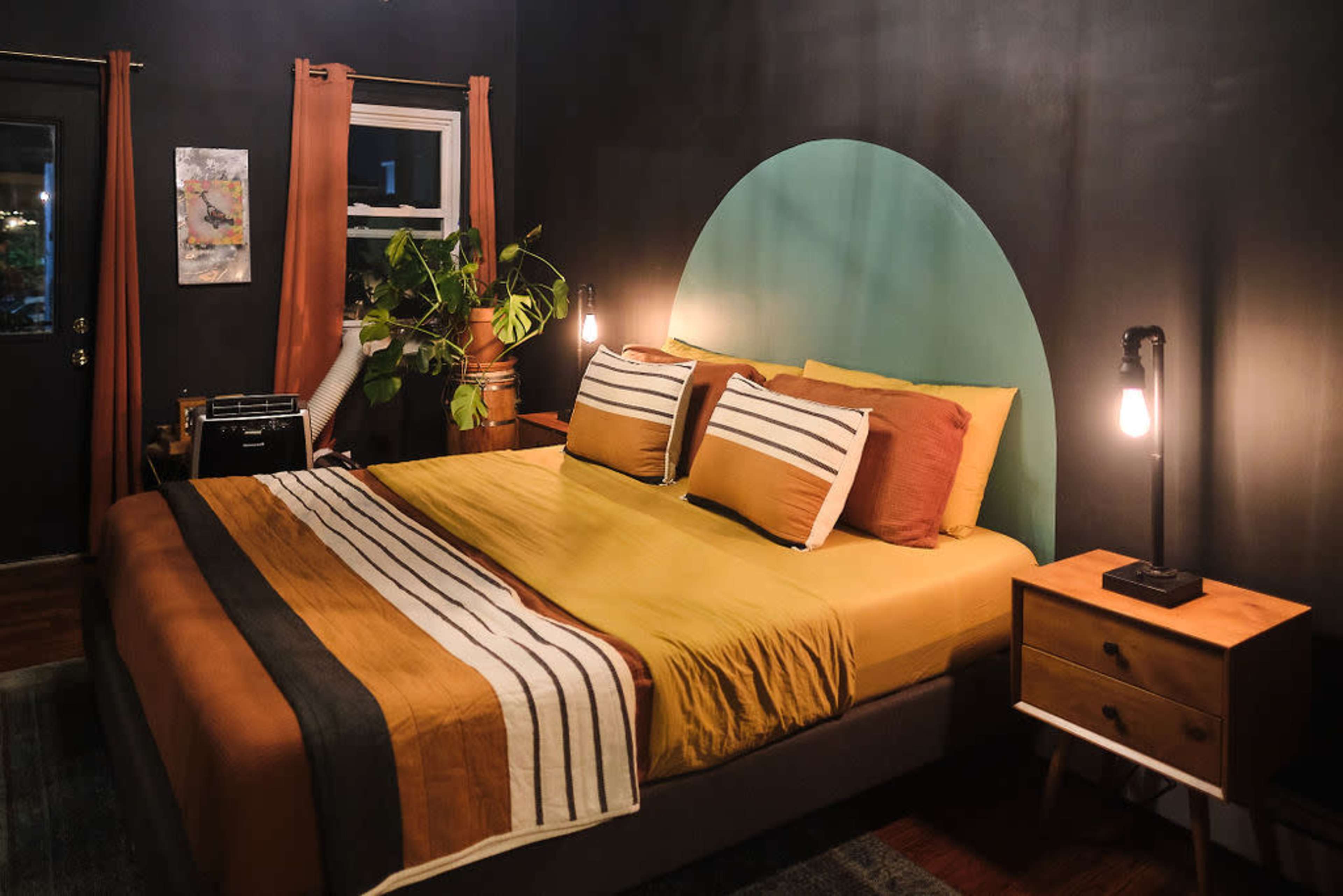 A bedroom with a large bed covered in yellow and brown bedding, green accent wall behind the headboard, and wooden furniture including bedside tables and a plant.