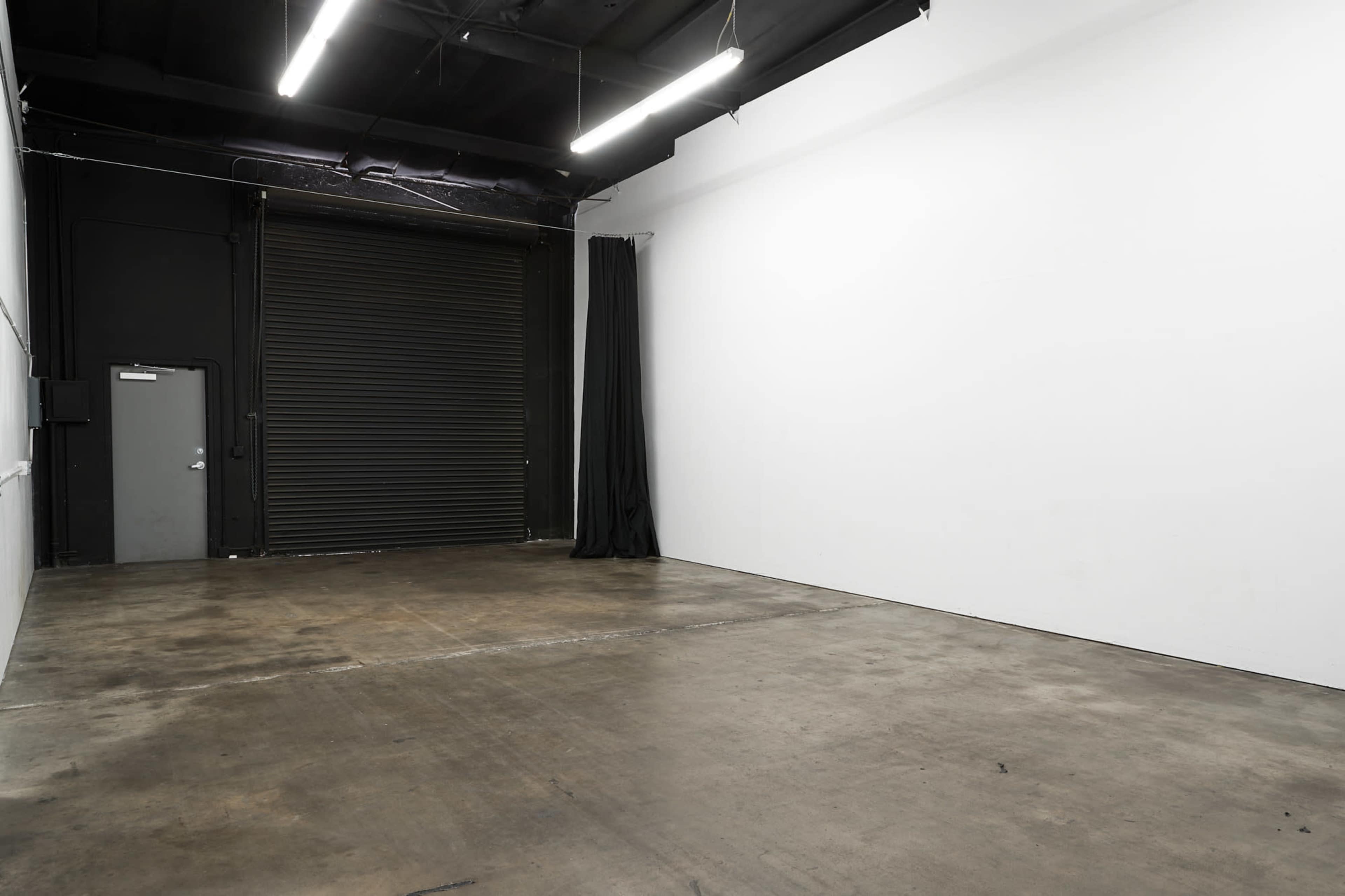 An empty industrial space with a concrete floor, a closed metal roll-up door, and a blank white wall.