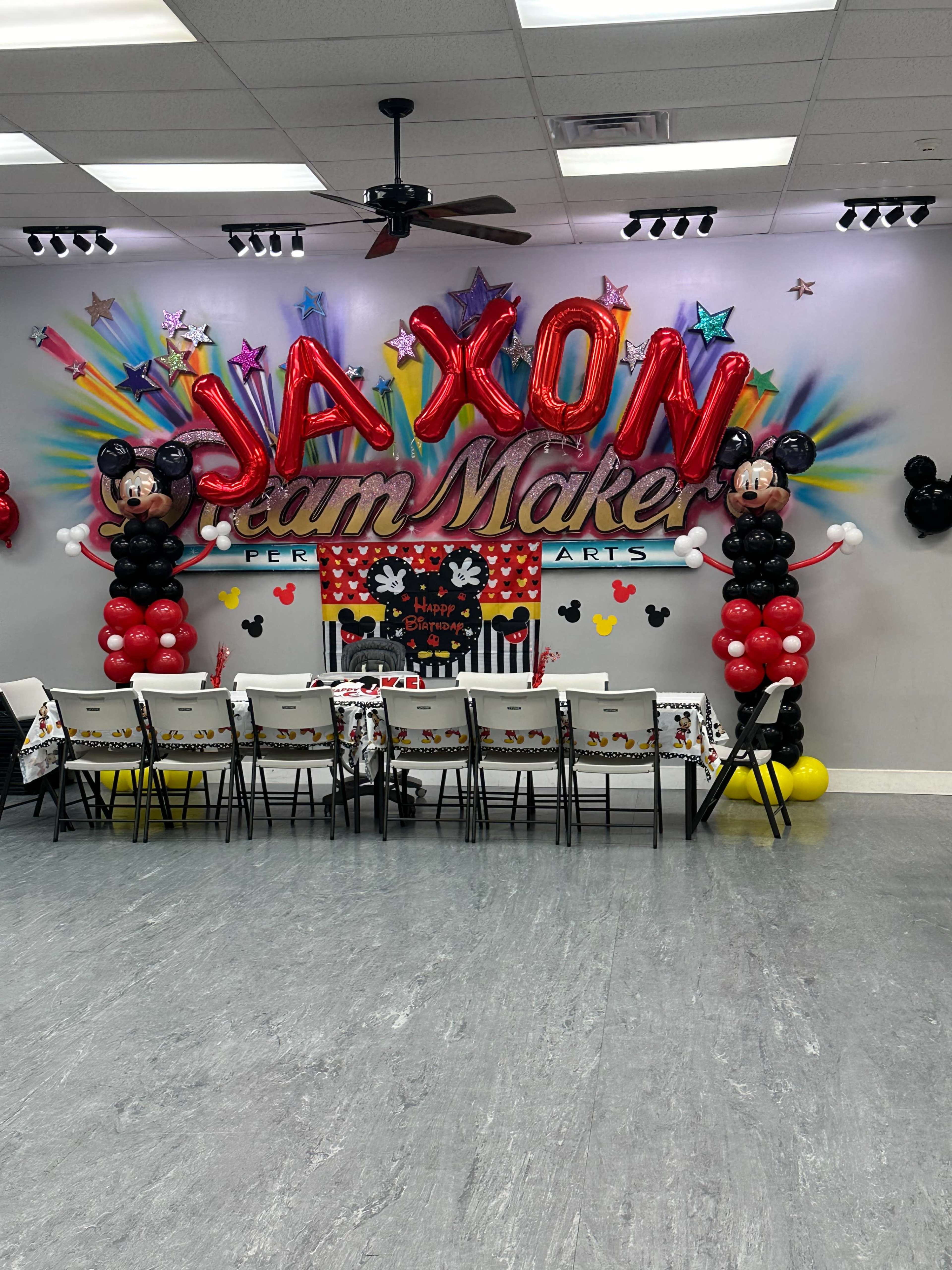 The image shows a colorful birthday party setup featuring large "JAXON" letters and Mickey Mouse balloon decorations, with a decorated table and chairs in front.