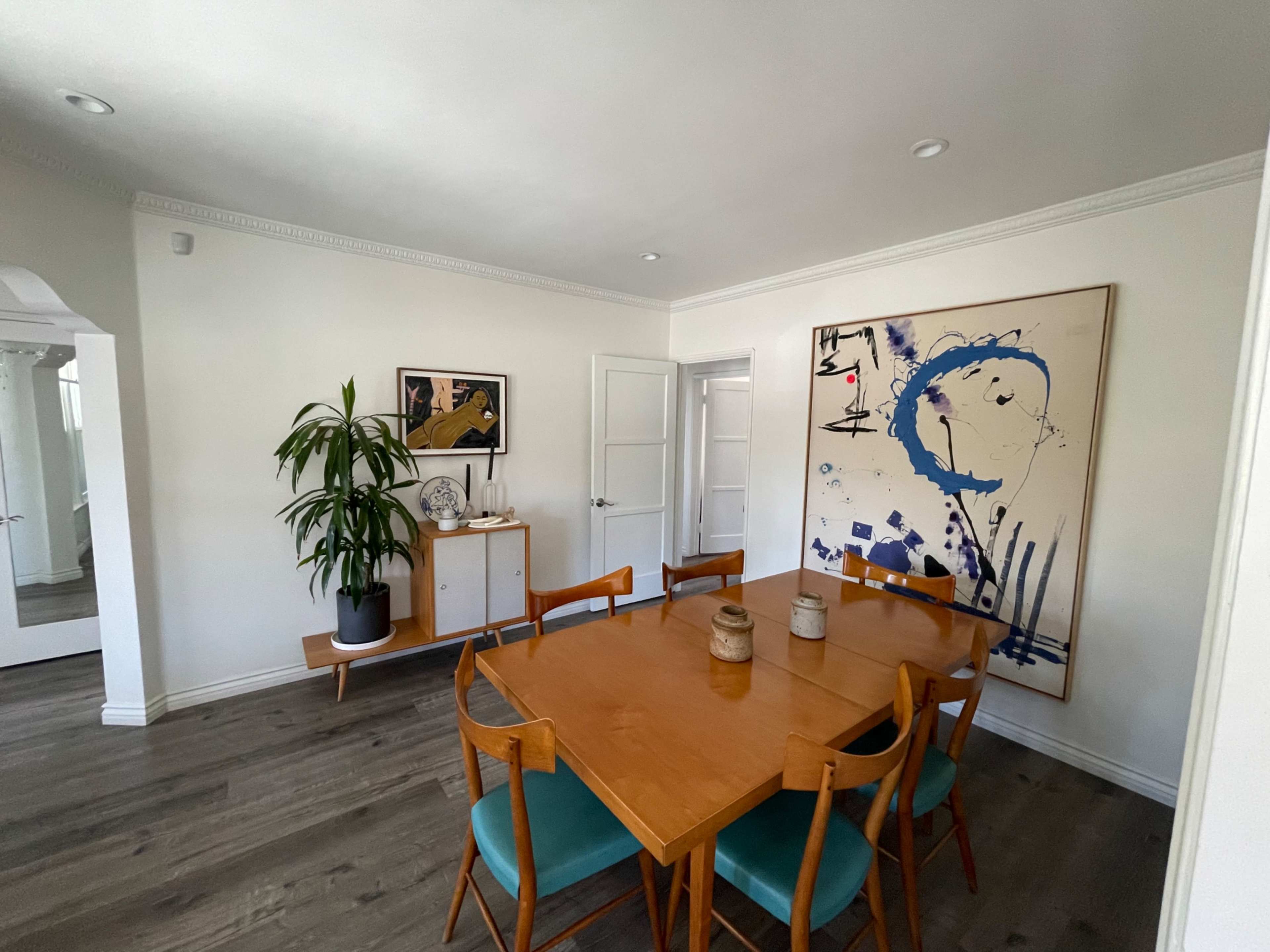 A dining area featuring a wooden table surrounded by chairs, a large abstract painting on the wall, and a potted plant in the corner.