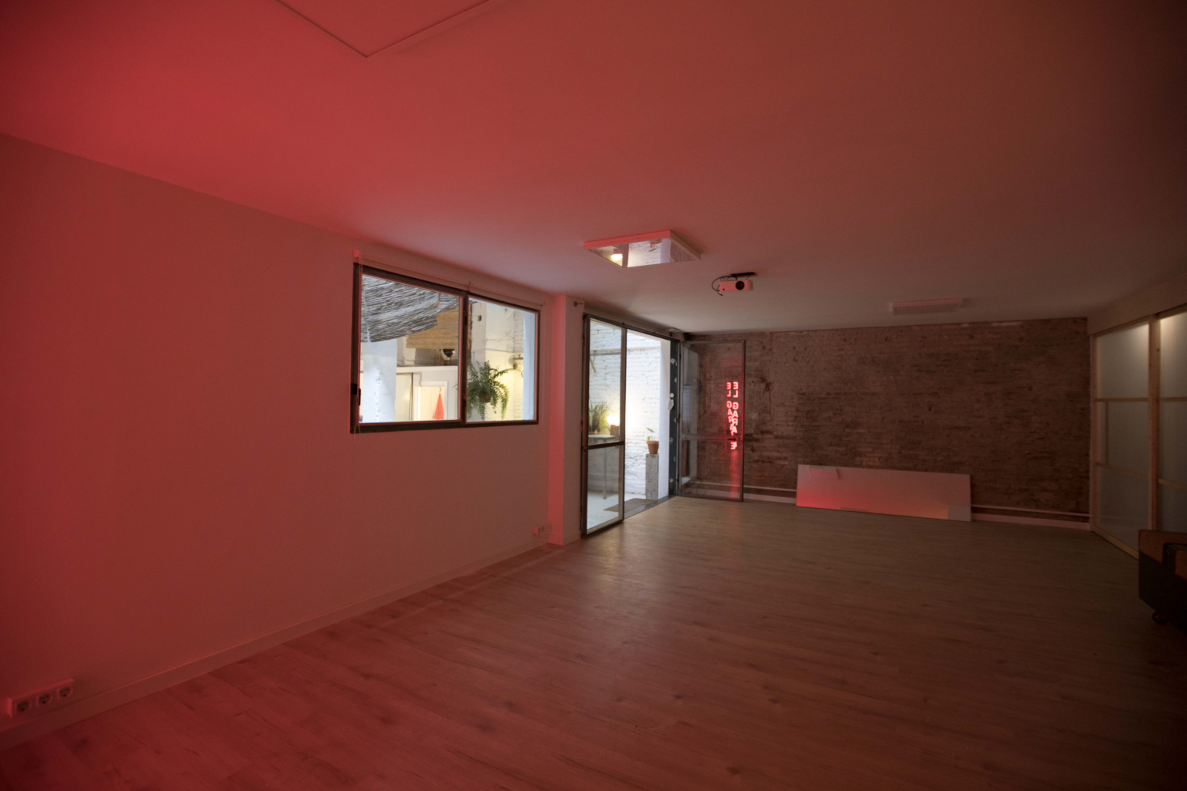 A sparsely furnished room with wooden flooring and red lighting, featuring large windows and a brick wall.