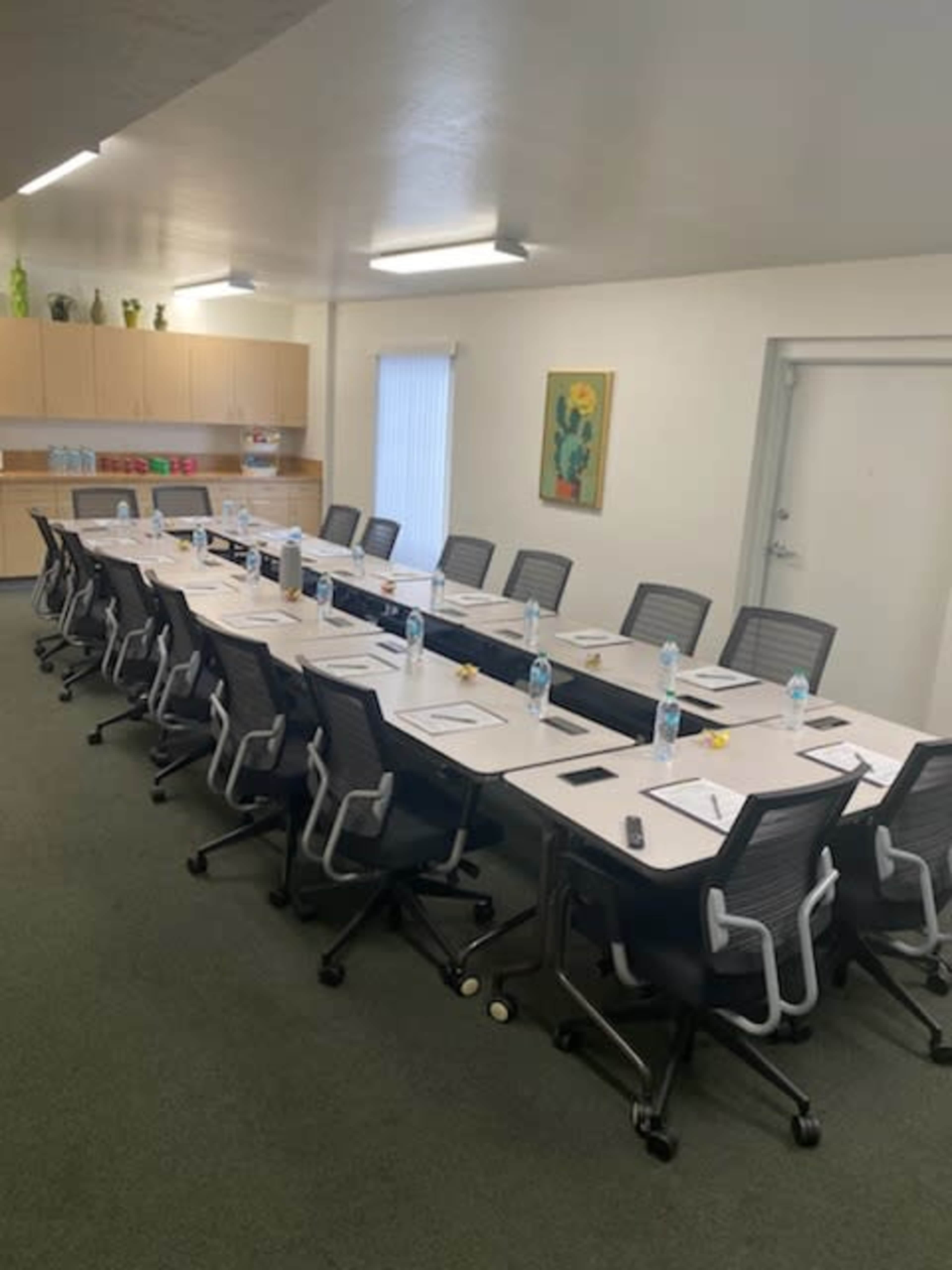 A long conference table is set up with chairs around it, water bottles, and notepads, in a well-lit meeting room featuring cabinets and a wall decoration.