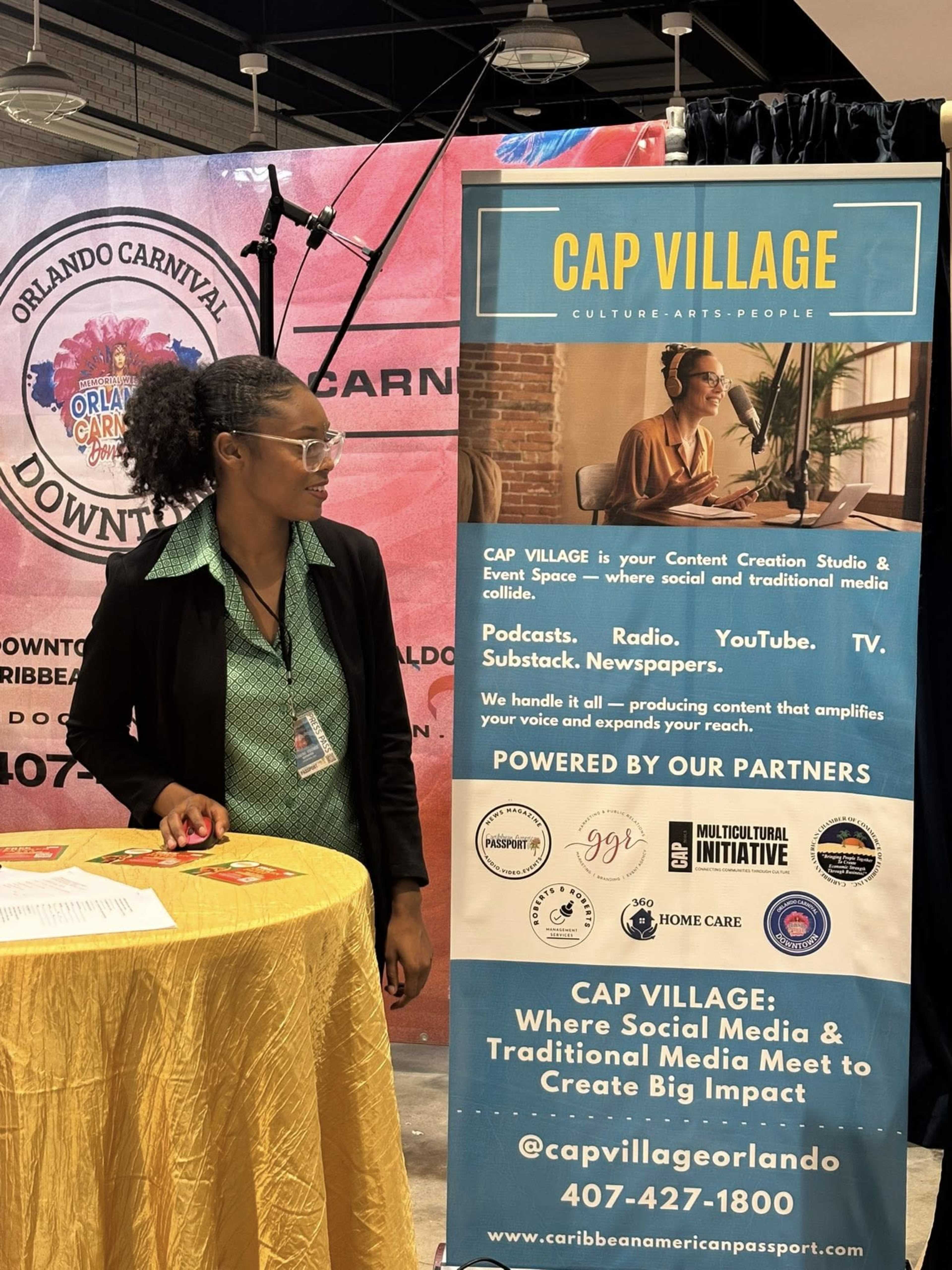 The image shows a promotional booth at an event with a banner that reads "CAP VILLAGE" and a table displaying information related to content creation and media engagement.