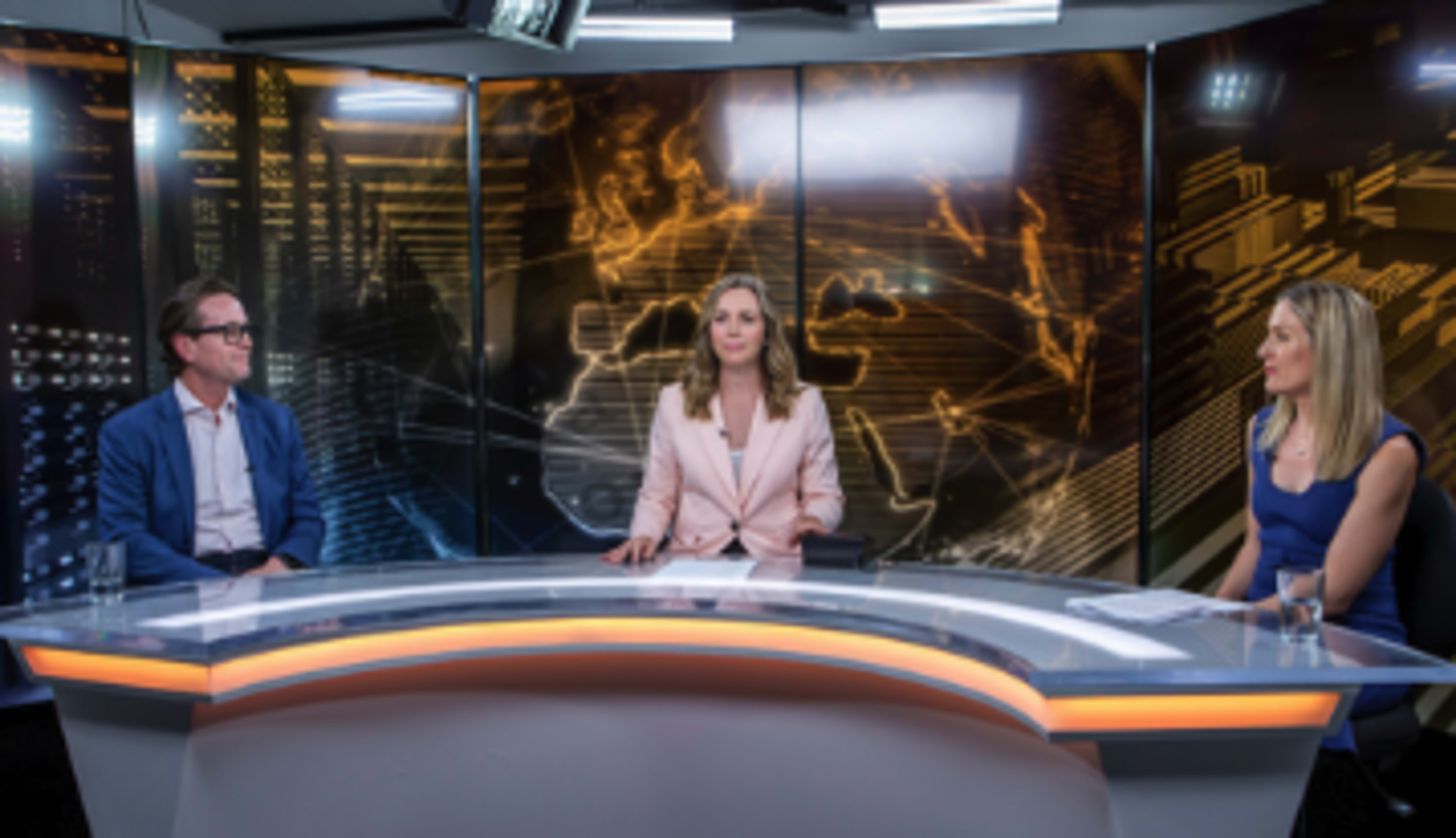 A news studio features three presenters seated at a curved table with a digital backdrop depicting a map and data graphics.