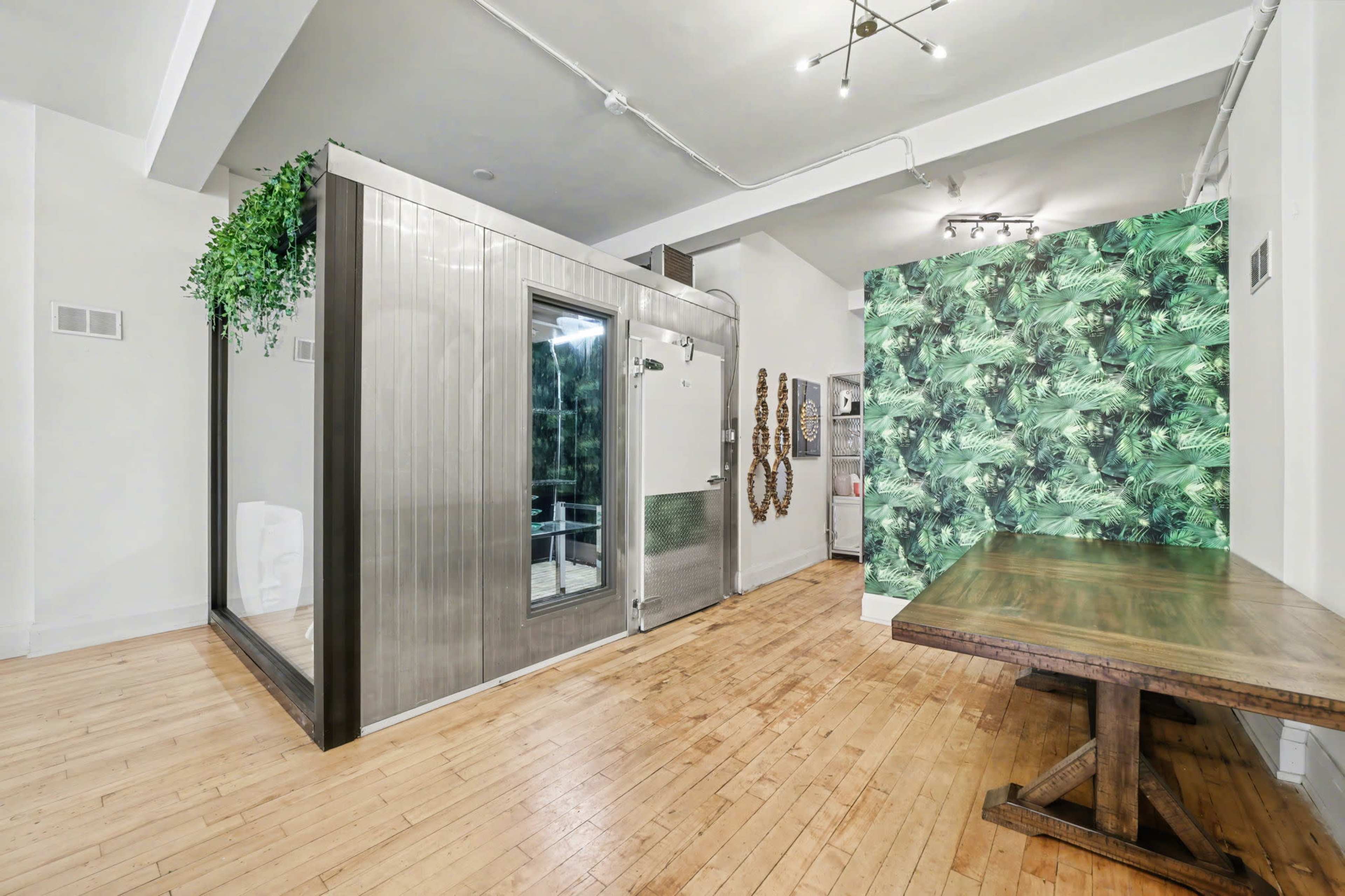 The image shows a modern interior space featuring a shower enclosed in a metallic frame, a wooden table, and a wall decorated with green tropical leaves.