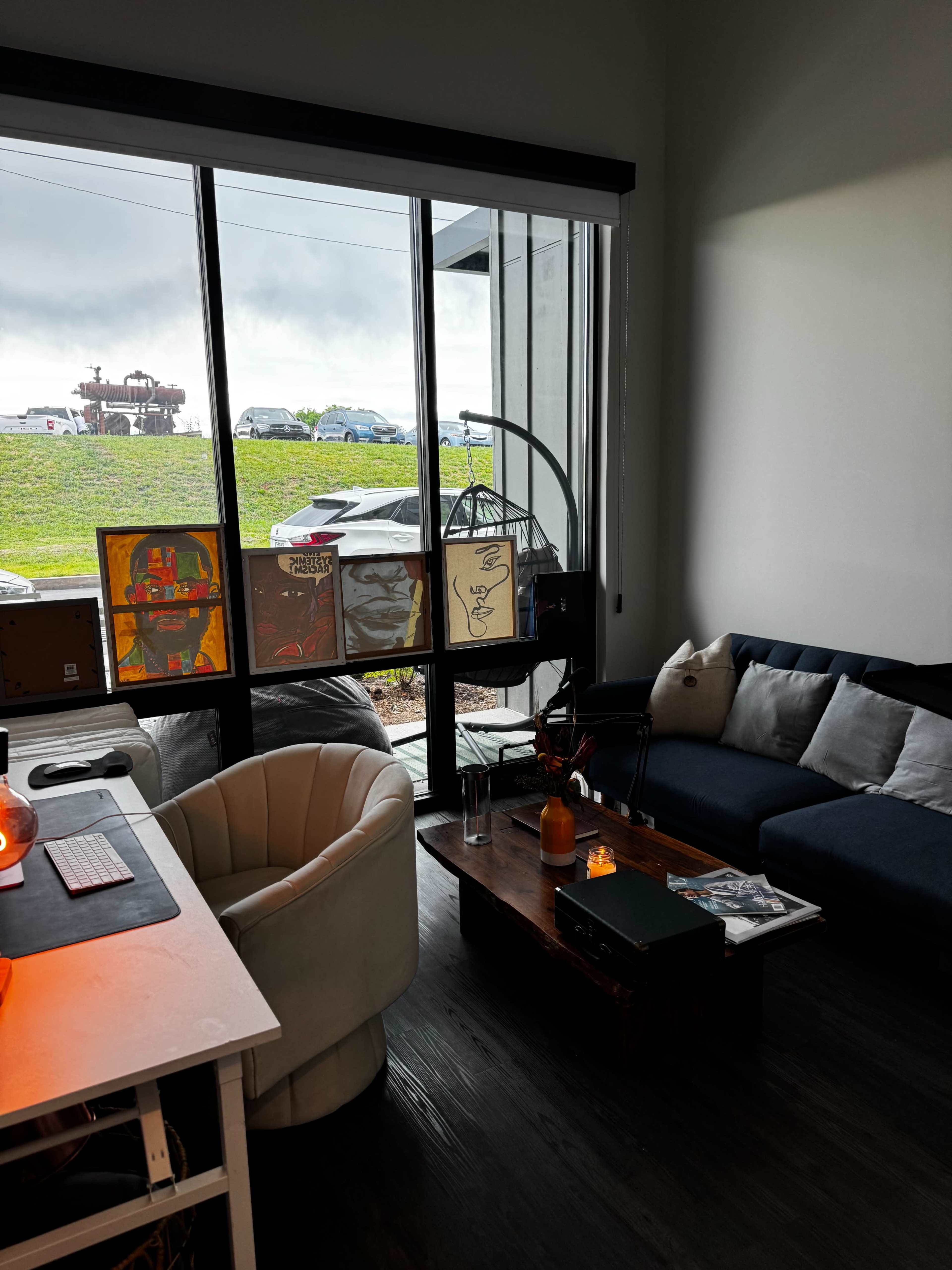 The image shows a modern office space featuring a white desk, a comfortable armchair, a dark sofa, and large windows revealing an outdoor view.