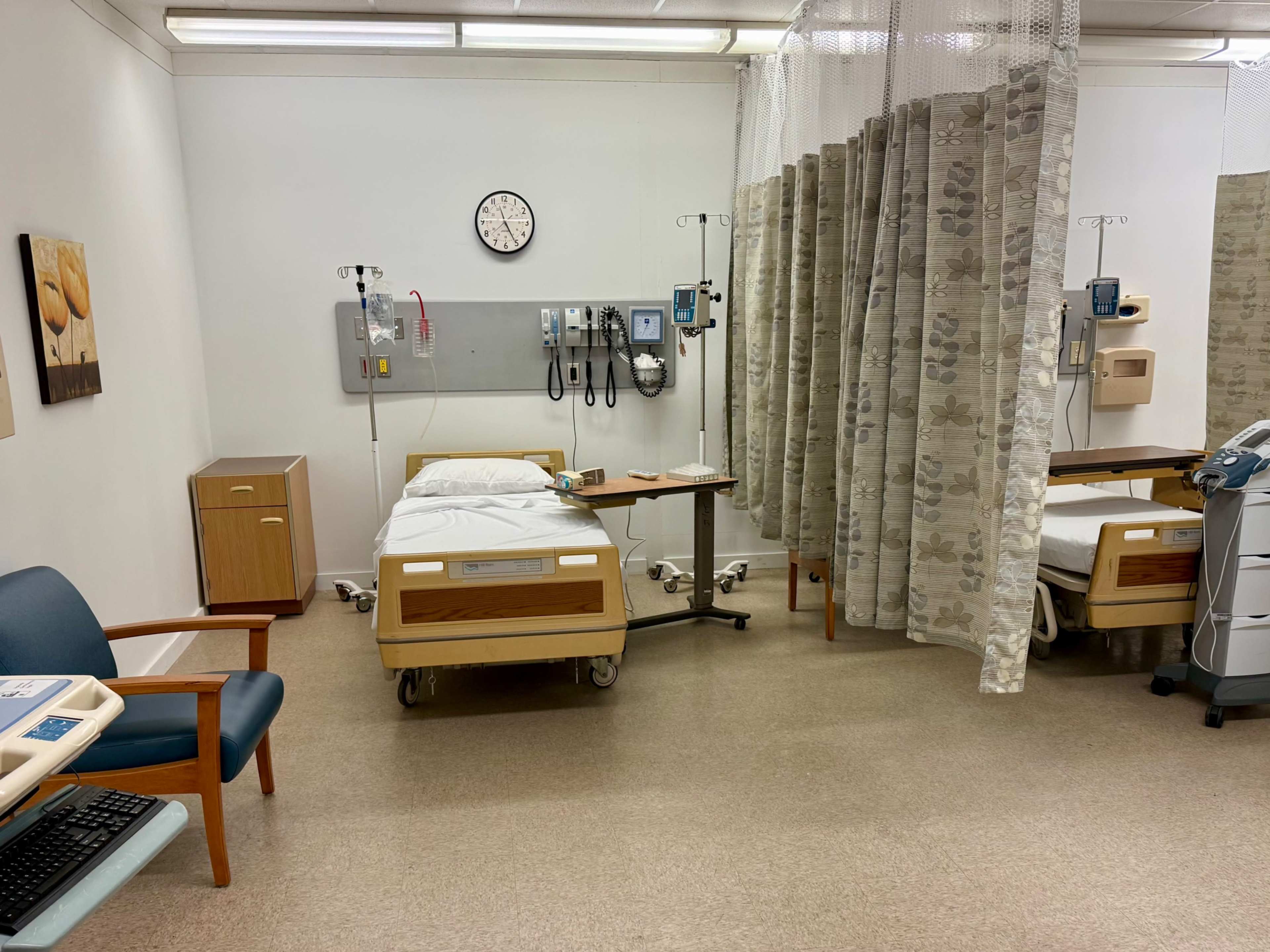 The image shows a hospital room with two empty beds, medical equipment on the walls, a clock, a small wooden cabinet, and privacy curtains.