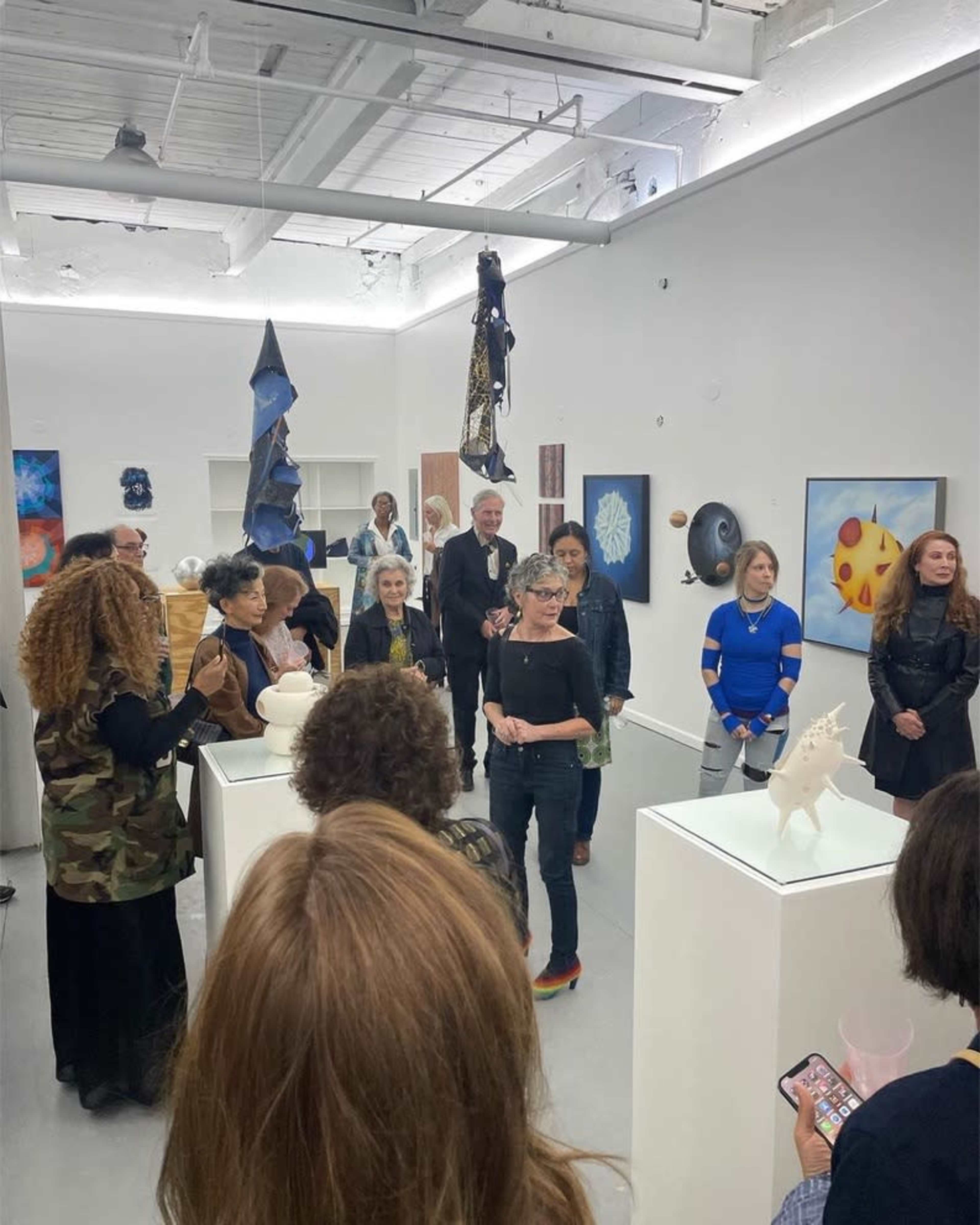A group of people is gathered in an art gallery, observing various artworks displayed on white pedestals and walls.