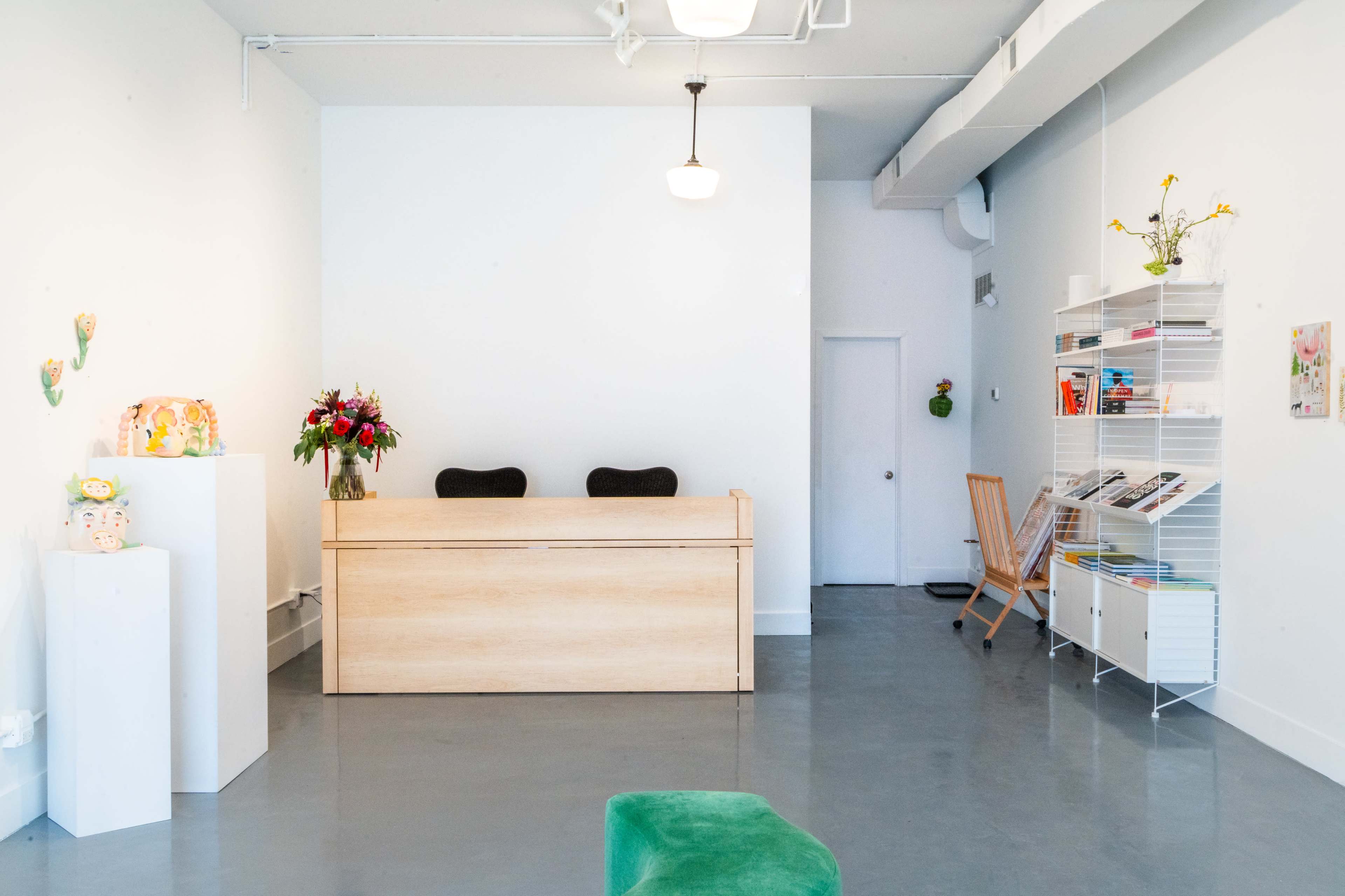 The image shows a minimalist reception area with a wooden front desk, a shelf filled with magazines, and a small waiting area featuring a wooden chair and a flower arrangement on display.