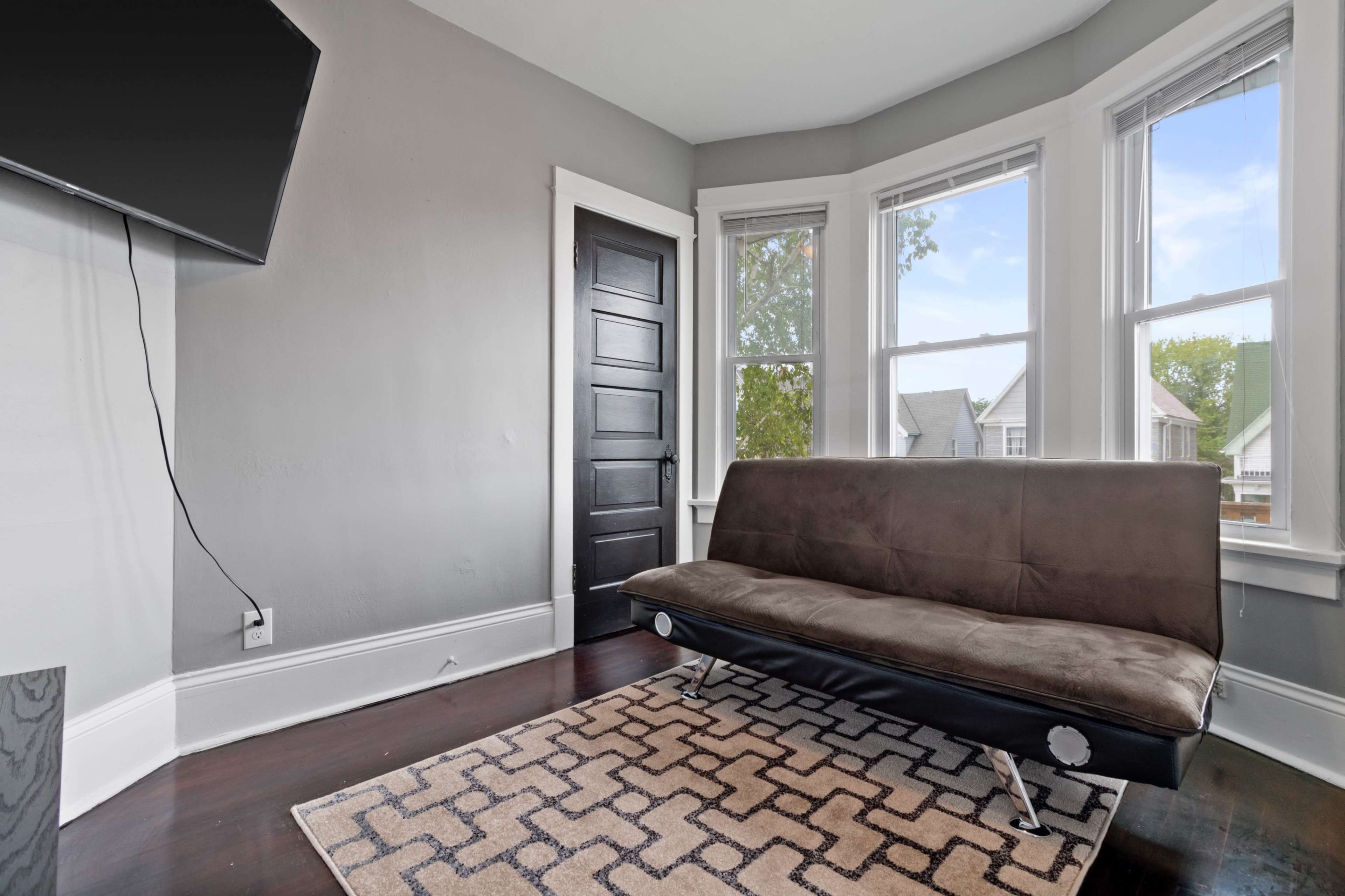 A small living room features a brown futon, a flat-screen TV mounted on the wall, and large windows allowing natural light to enter.